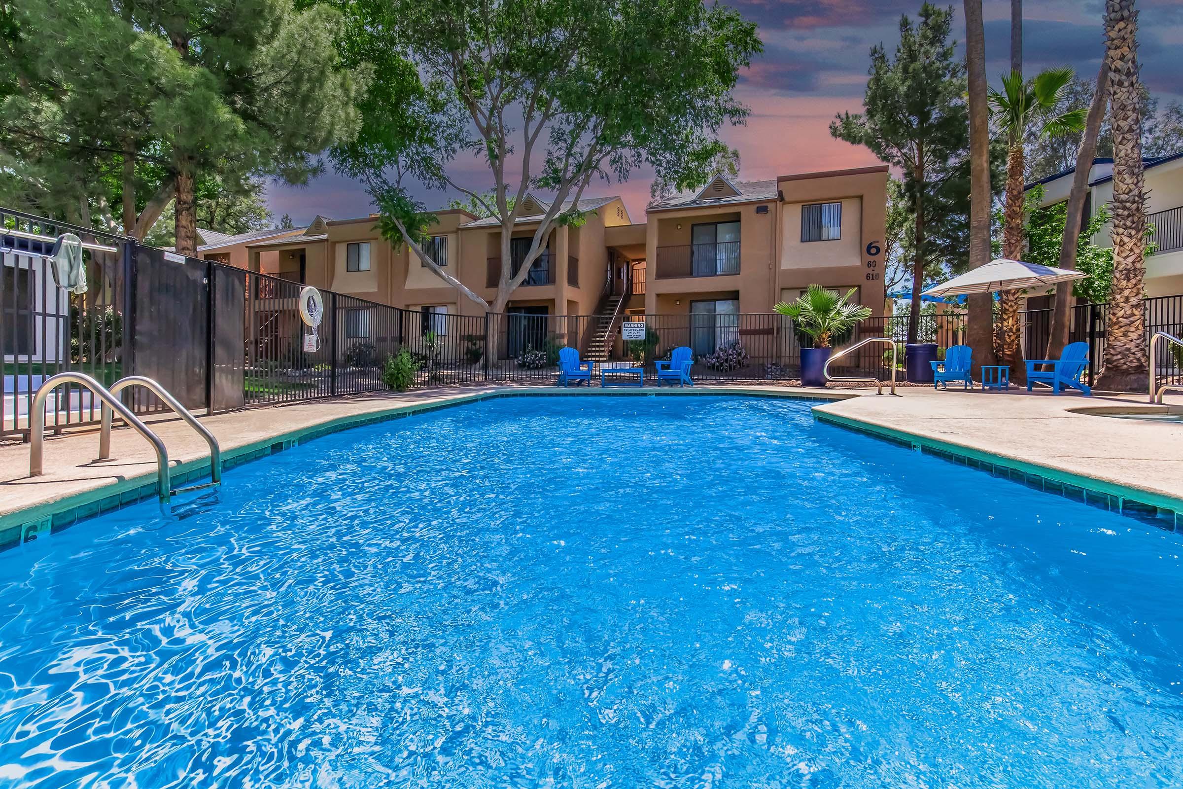A refreshing swimming pool surrounded by palm trees and lounge chairs, with apartment buildings in the background. The sky features a colorful sunset. The area is enclosed with a fence, creating a relaxing atmosphere for residents.