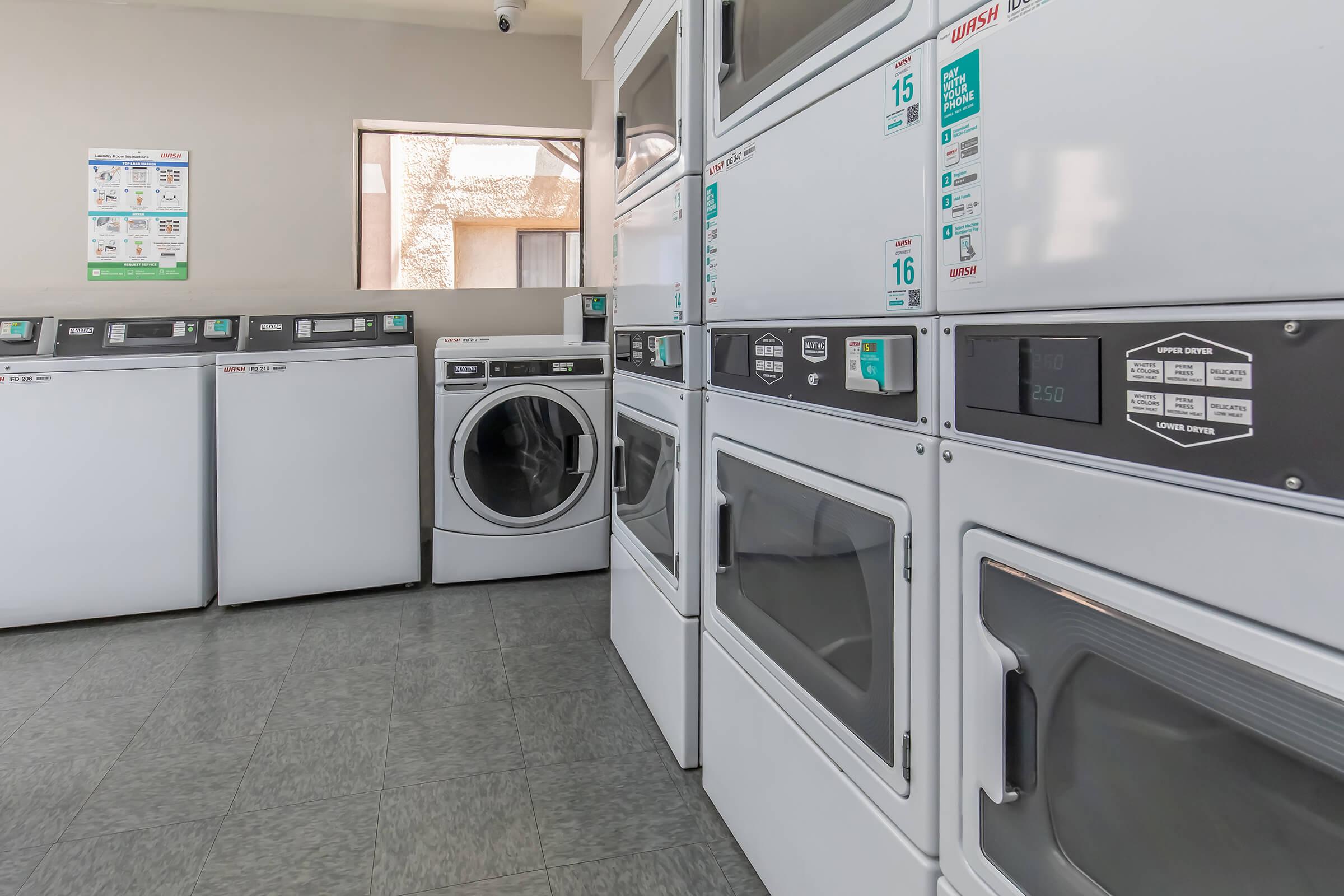 A clean and well-lit laundry room featuring several stacked washing machines and dryers. The machines have digital displays and buttons for user convenience. A window offers natural light, and informational posters about laundry usage are visible on the wall. The floor is tiled with a light gray pattern.