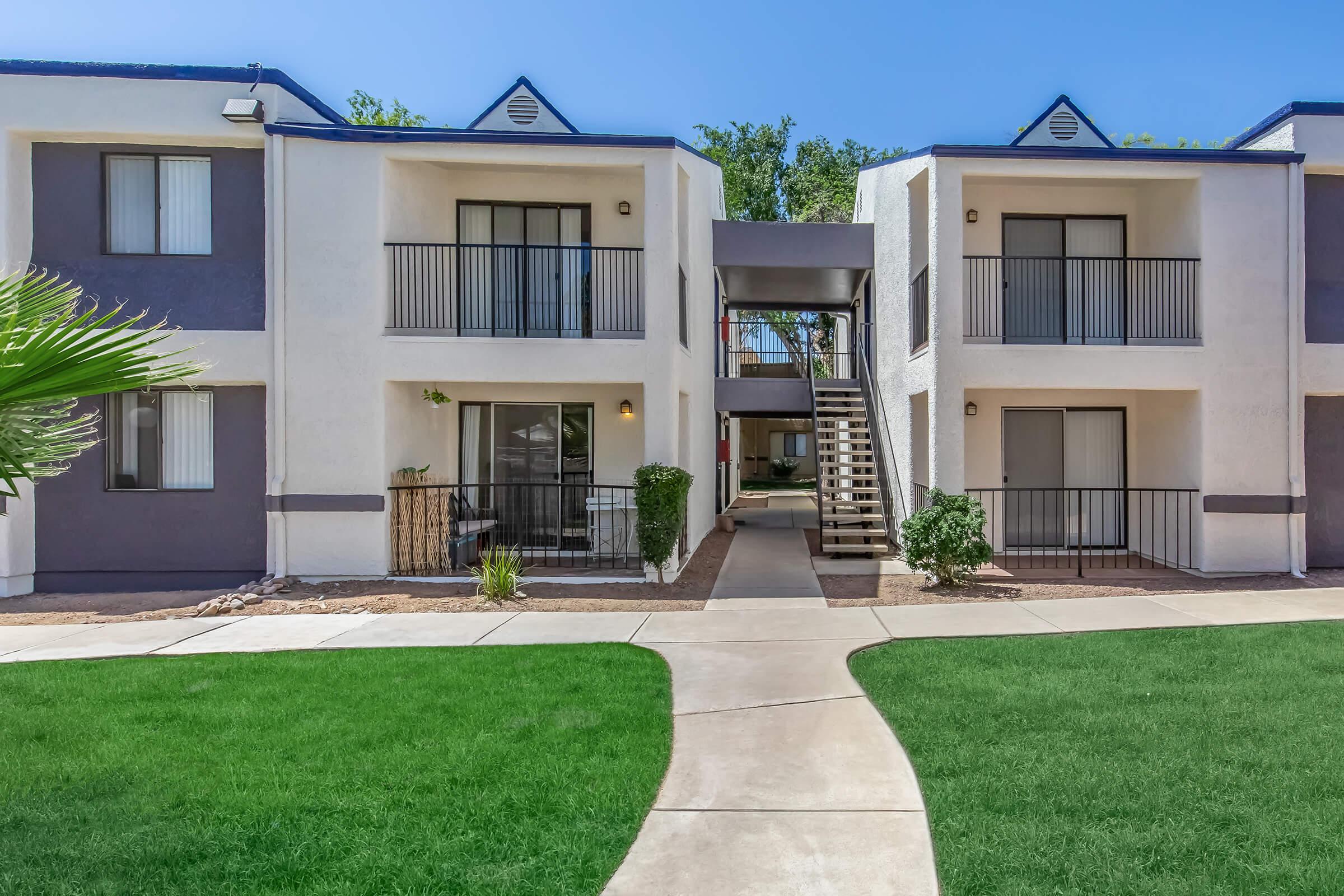 A well-maintained apartment complex featuring two-story buildings with balconies, surrounded by green lawn and landscaped areas. A pathway leads through the center, connecting the units, and stairs provide access to the upper floors. Clear blue sky overhead adds to the inviting atmosphere.