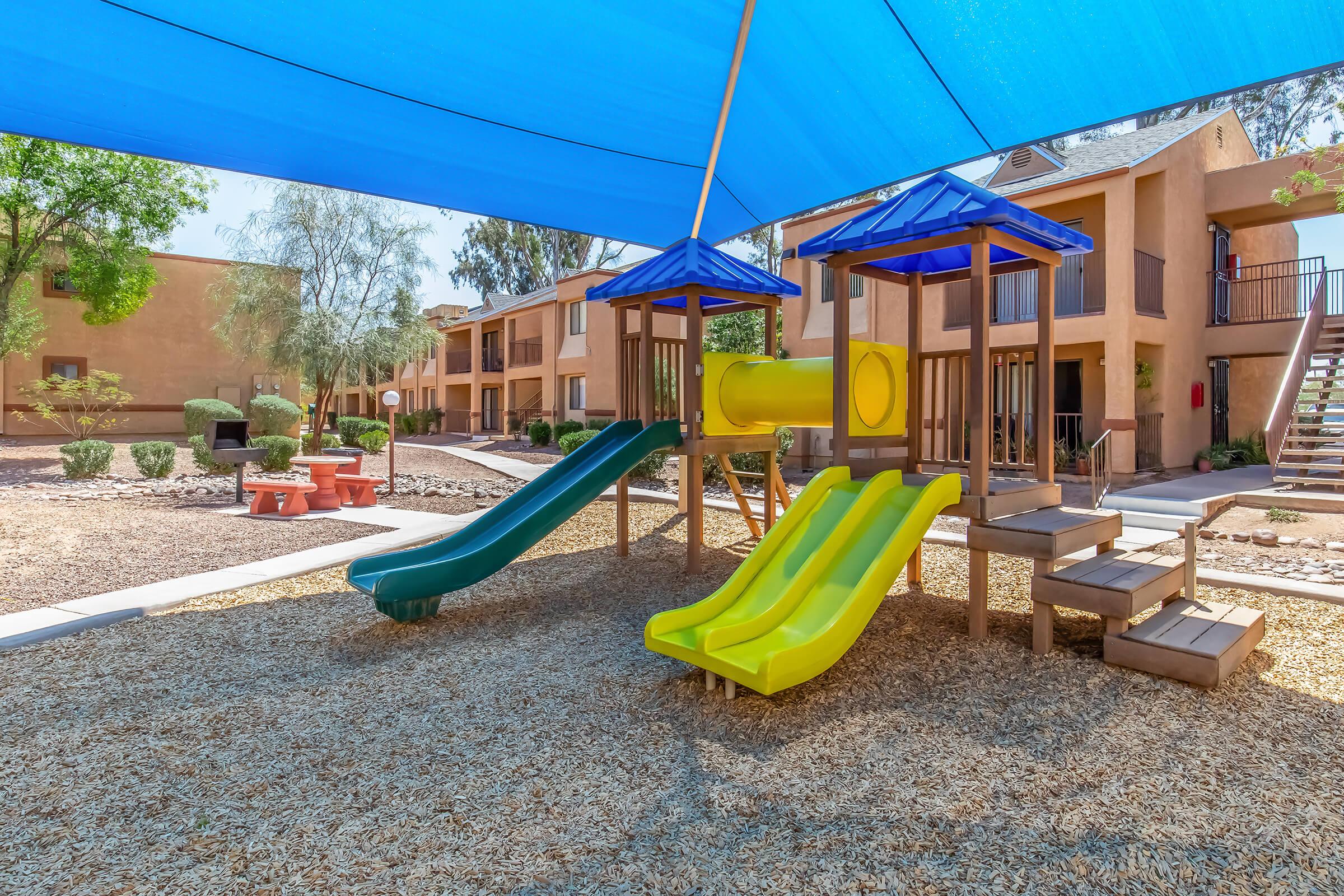 Playground area with two slides, one green and one yellow, under a blue shade structure. Surrounding the playground are landscaped areas with shrubs and a picnic table nearby. In the background, there are two-story apartment buildings with a clear sky above.