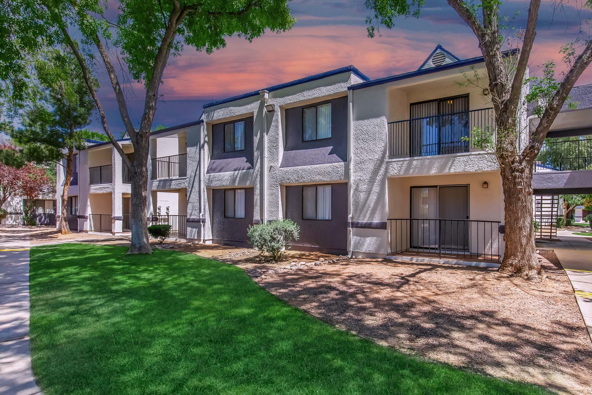 A view of a two-story apartment building surrounded by green grass and trees. The building features balconies and a well-maintained landscape with gravel areas. A colorful sky is visible in the background, suggesting sunset or sunrise lighting.