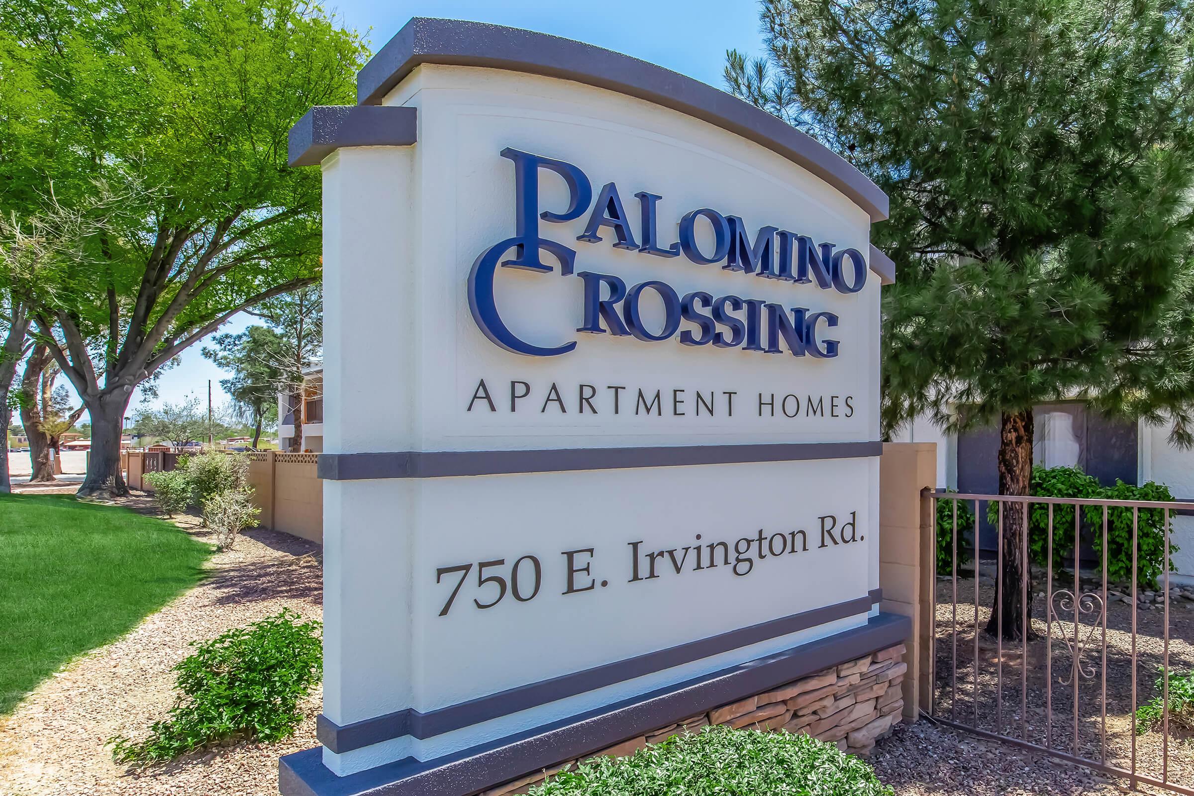 Sign for Palomino Crossing Apartment Homes located at 750 E. Irvington Rd. The sign is prominently displayed and surrounded by greenery, including trees and shrubs, providing a welcoming entrance to the apartment community.