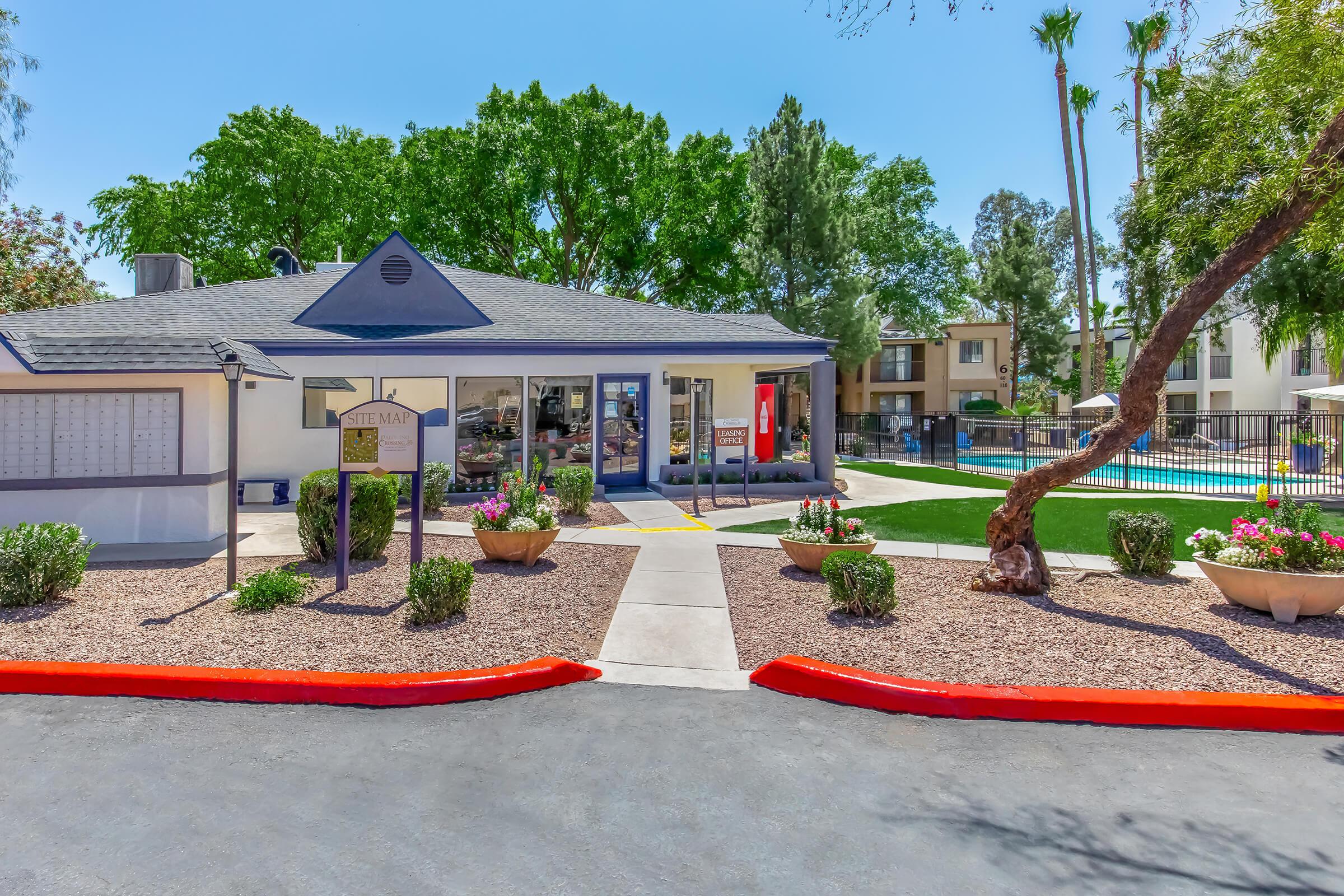 A view of an apartment complex entrance featuring a spacious pathway, landscaped gardens with potted plants, and a swimming pool in the background. The building has a modern design with large windows and is surrounded by trees, providing a welcoming atmosphere.
