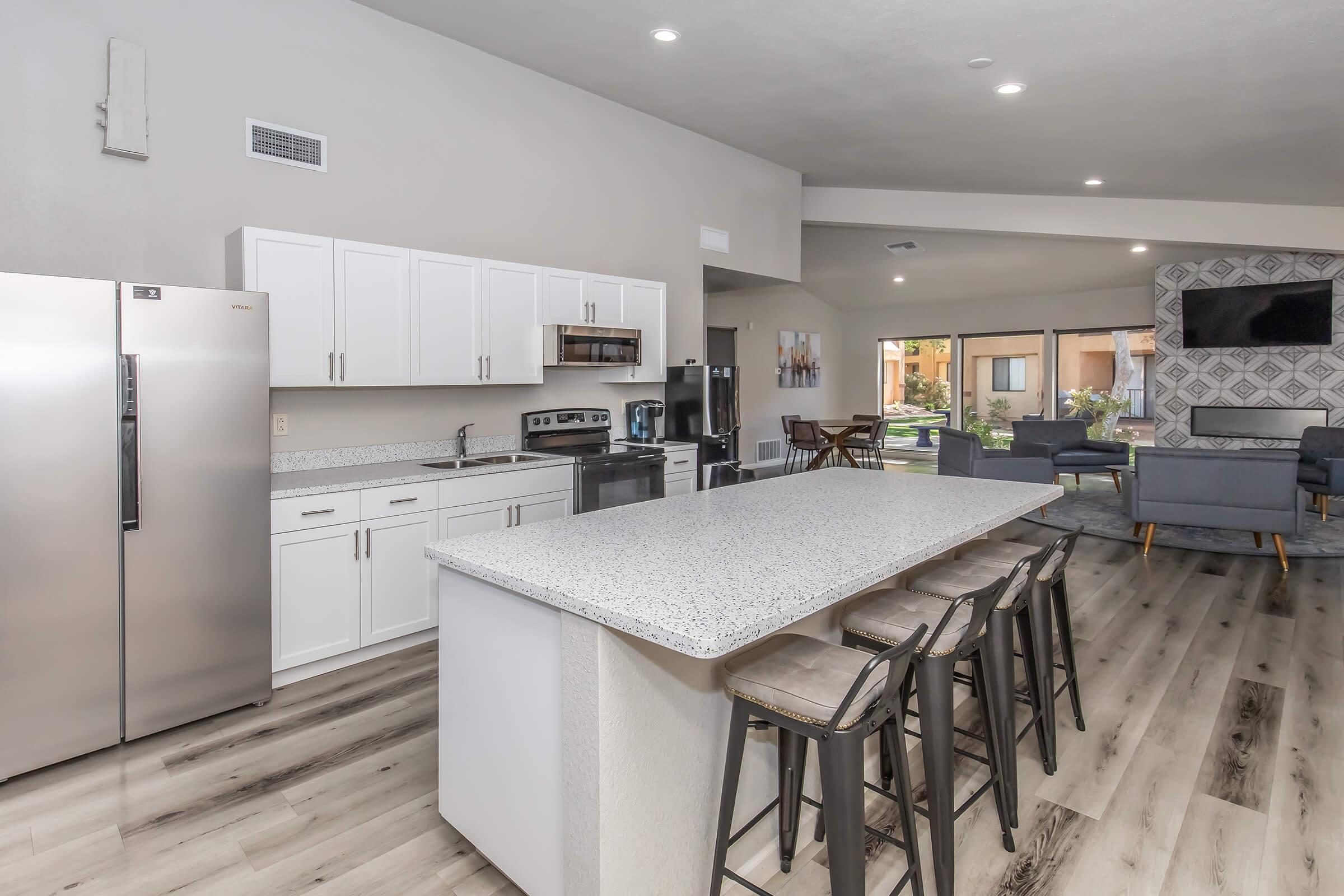 Modern kitchen with stainless steel appliances, including a refrigerator and stove, white cabinets, and a large island with barstools. The open space features gray wood flooring and a cozy living area visible in the background, with large windows letting in natural light.