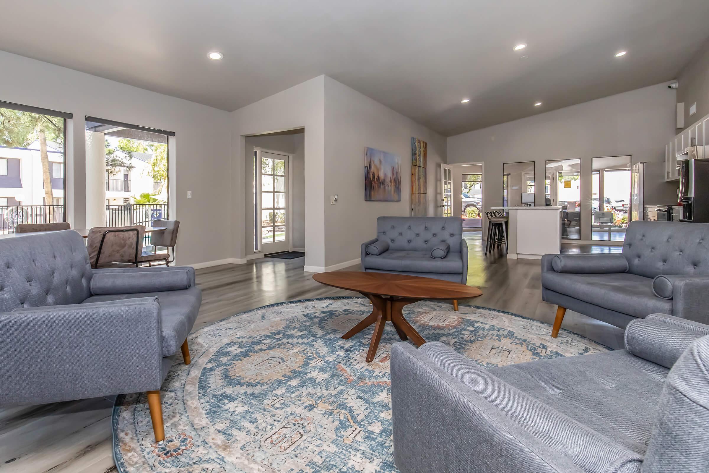 A modern living room featuring gray upholstered armchairs and a round wooden coffee table on a patterned area rug. Large windows allow natural light to enter, and there's a view of the outdoors. The walls are painted light gray, and artwork adorns one side, creating a spacious and welcoming atmosphere.