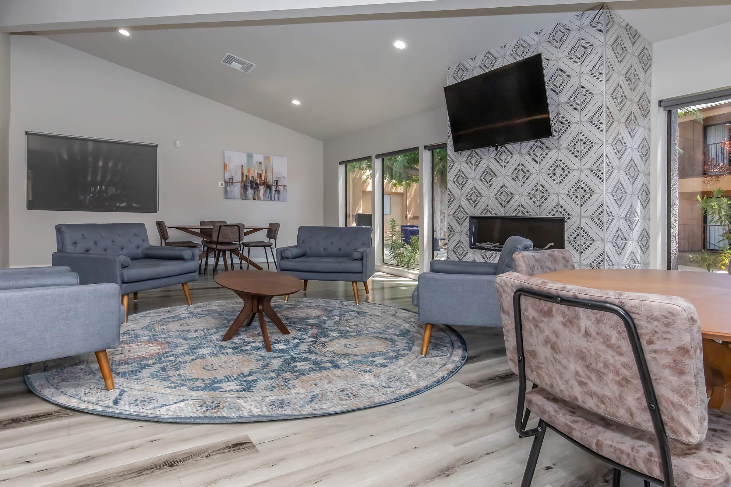 A modern living space featuring gray upholstered chairs around a circular wooden table on a decorative rug. A cozy fireplace and large windows provide natural light, while a textured wall adds visual interest. A flat-screen TV is mounted on the wall, enhancing the inviting atmosphere.