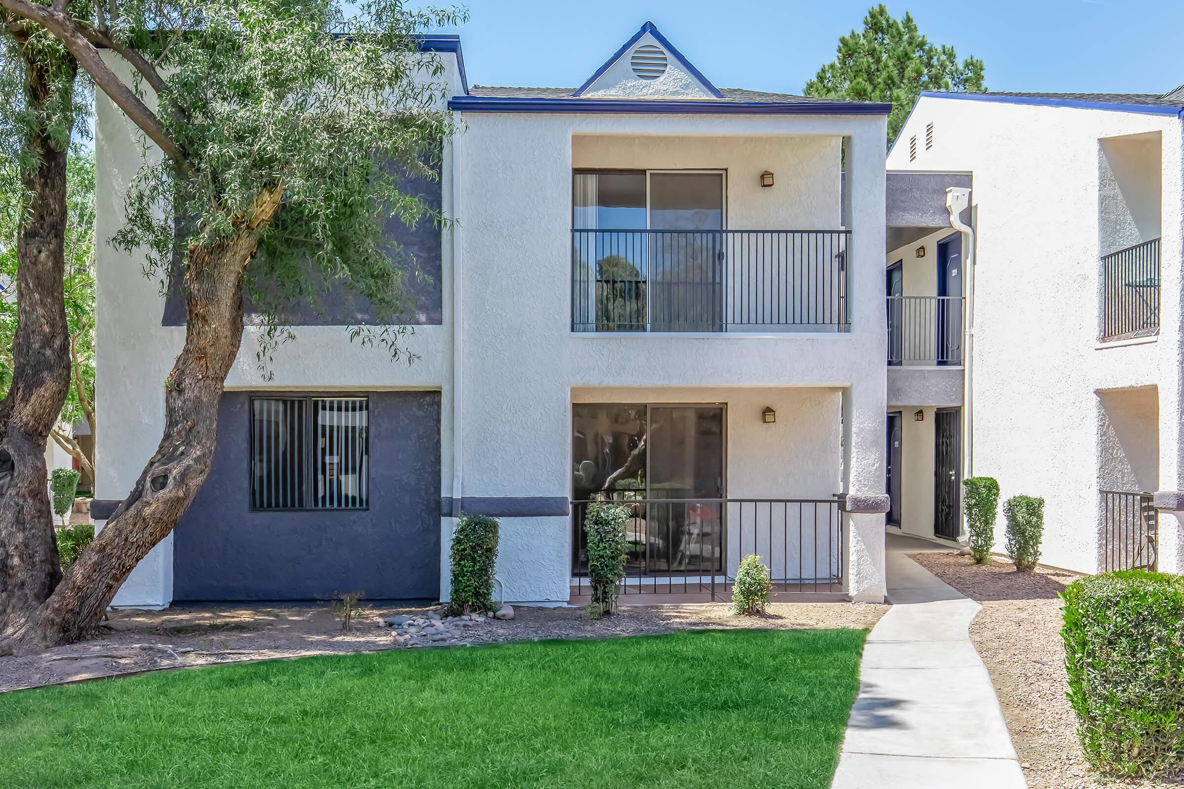 Two-story apartment building with a white and light gray exterior. It features a balcony on the second floor, large windows, and landscaped greenery surrounding the building. A pathway leads to the entrance from a lawn area. Trees are visible nearby.