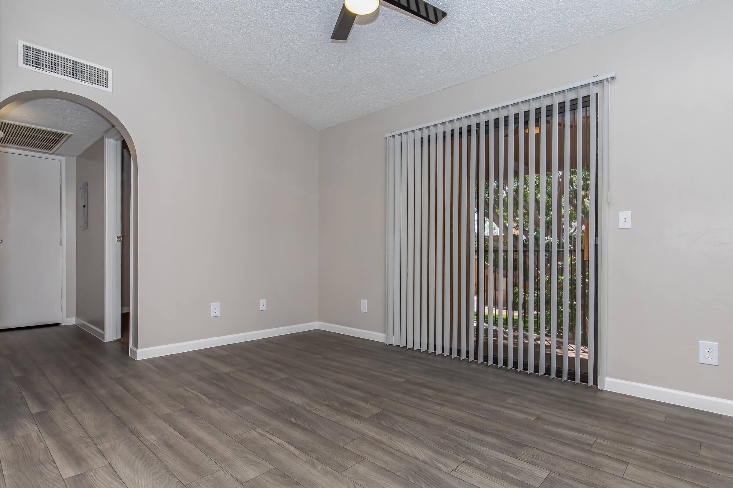 A spacious interior room with gray walls and wood-look flooring. There is an archway leading to another area, a ceiling fan overhead, and a large sliding glass door with vertical blinds that opens to an outdoor view.