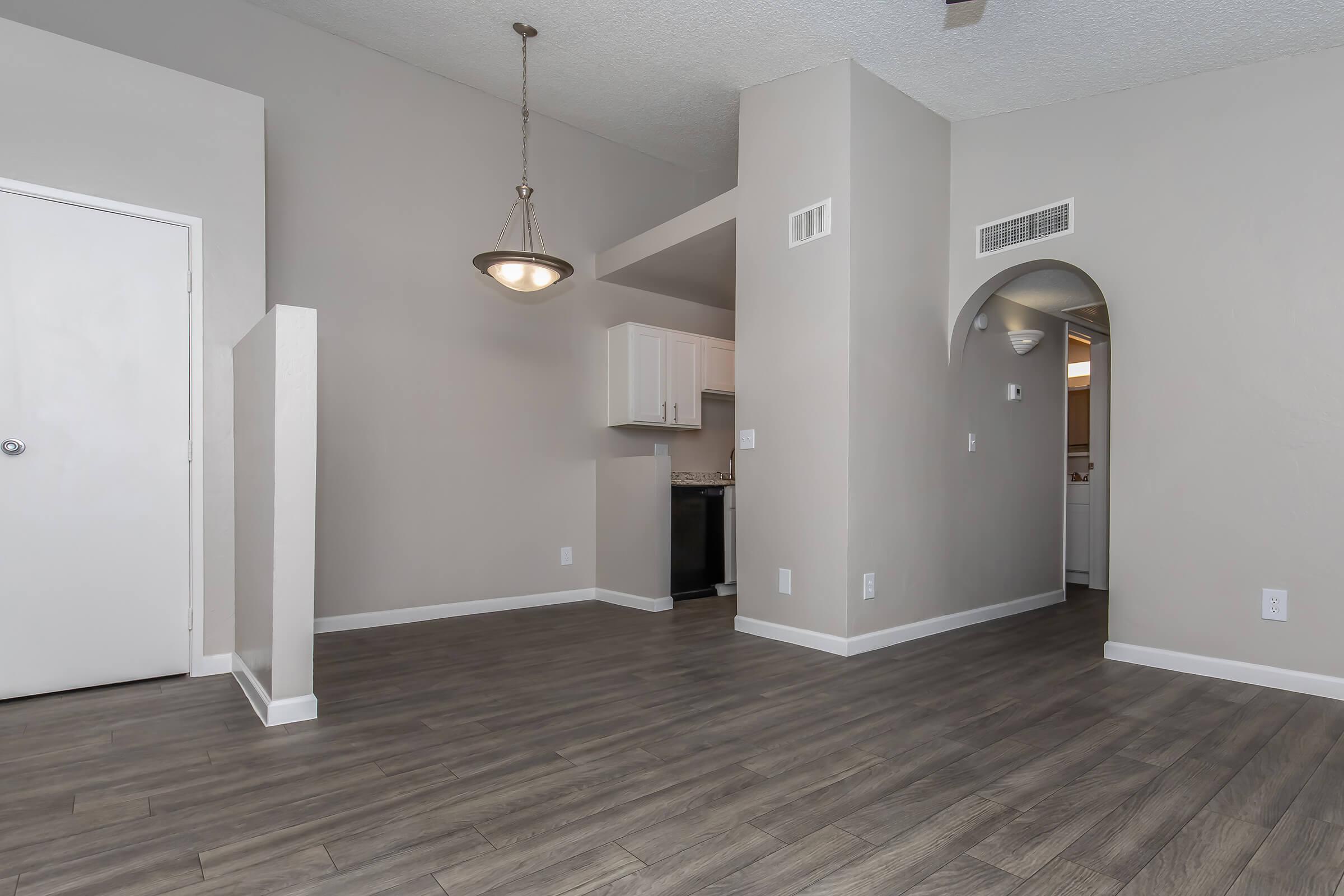 Spacious interior of an apartment featuring beige walls, laminate flooring, and a modern light fixture. The layout includes a doorway, archway, and a kitchen area visible in the background, with white cabinetry and appliances. Natural light enhances the inviting atmosphere.
