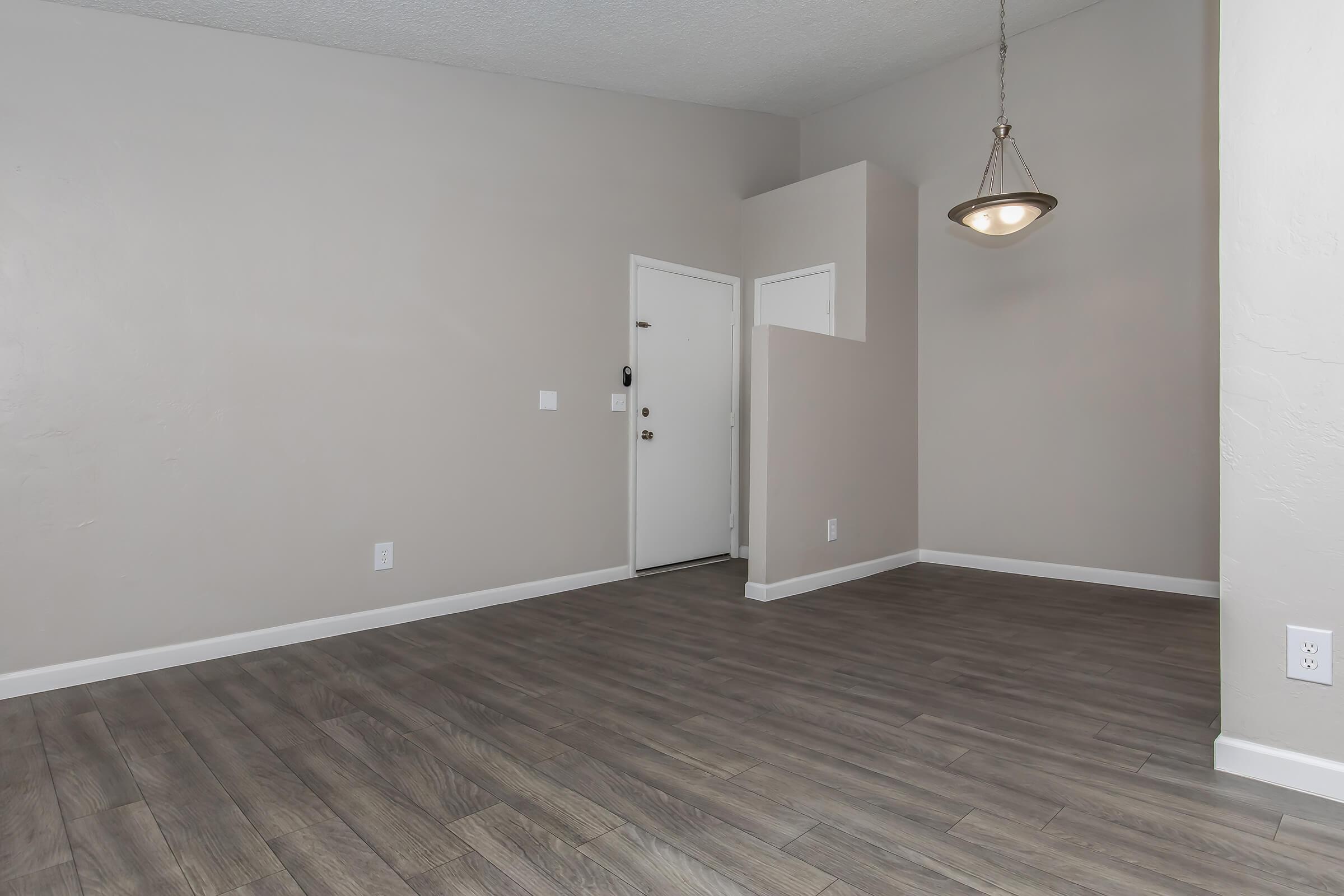 A spacious interior of an empty room with light gray walls and new laminate flooring. There's a doorway with a white door on the left and a small entrance area. A pendant light hangs from the ceiling, adding a modern touch to the otherwise minimalistic space. The room is well-lit and inviting.
