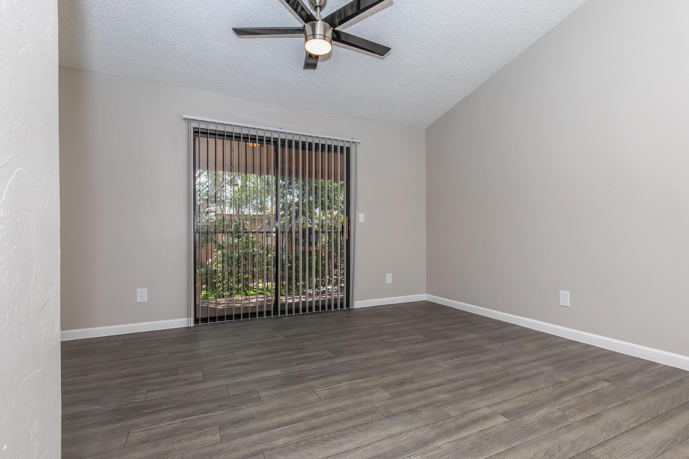 A spacious, well-lit room featuring a ceiling fan, light-colored walls, and modern vinyl flooring. Large glass sliding doors with blinds provide access to an outdoor view, illuminating the area with natural light. The space is empty, creating a blank canvas for furnishings and decoration.