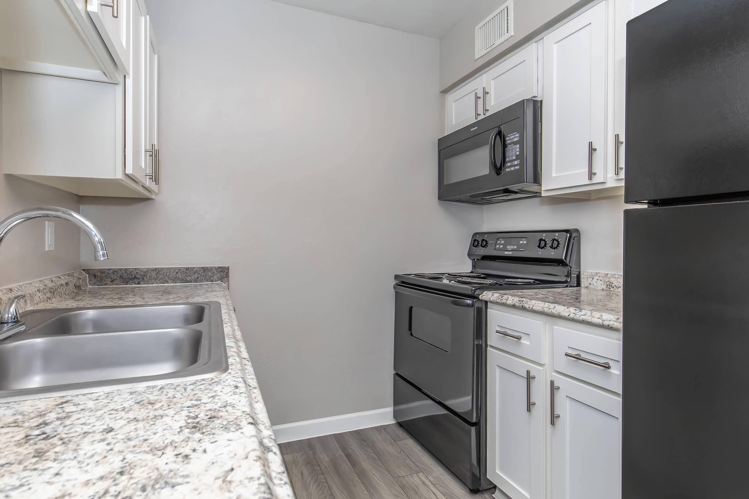 A modern kitchen featuring light-colored cabinets, a granite countertop, a double sink, a black stove, and a microwave. The walls are painted a soft gray, and the flooring is a light wood grain, creating a clean and contemporary look.