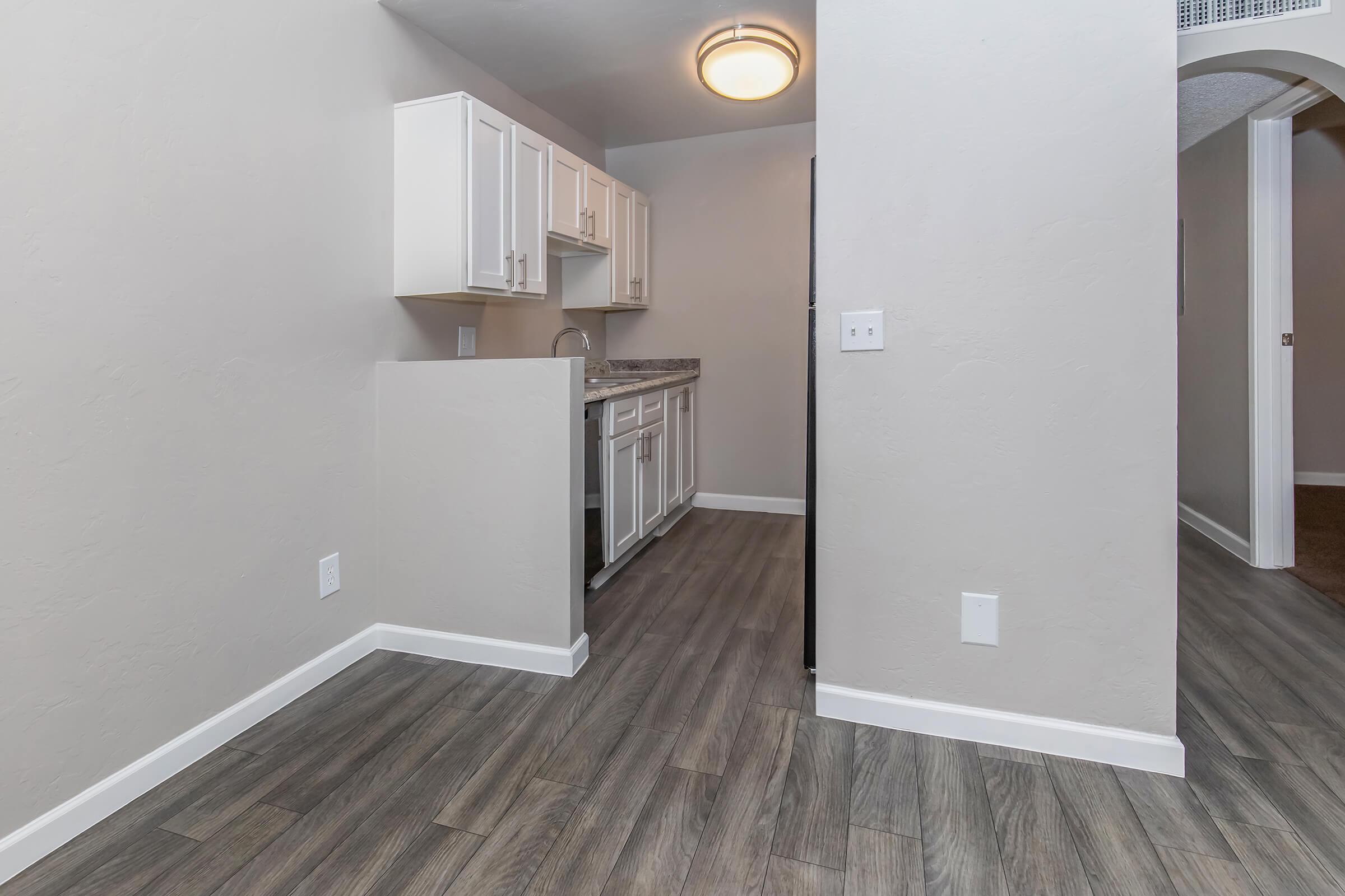A residential kitchen area featuring white cabinets, a granite countertop, and a modern light fixture. The flooring is a wood-like texture, and the walls are a neutral shade. Part of the open layout is visible, leading to another room in the background.