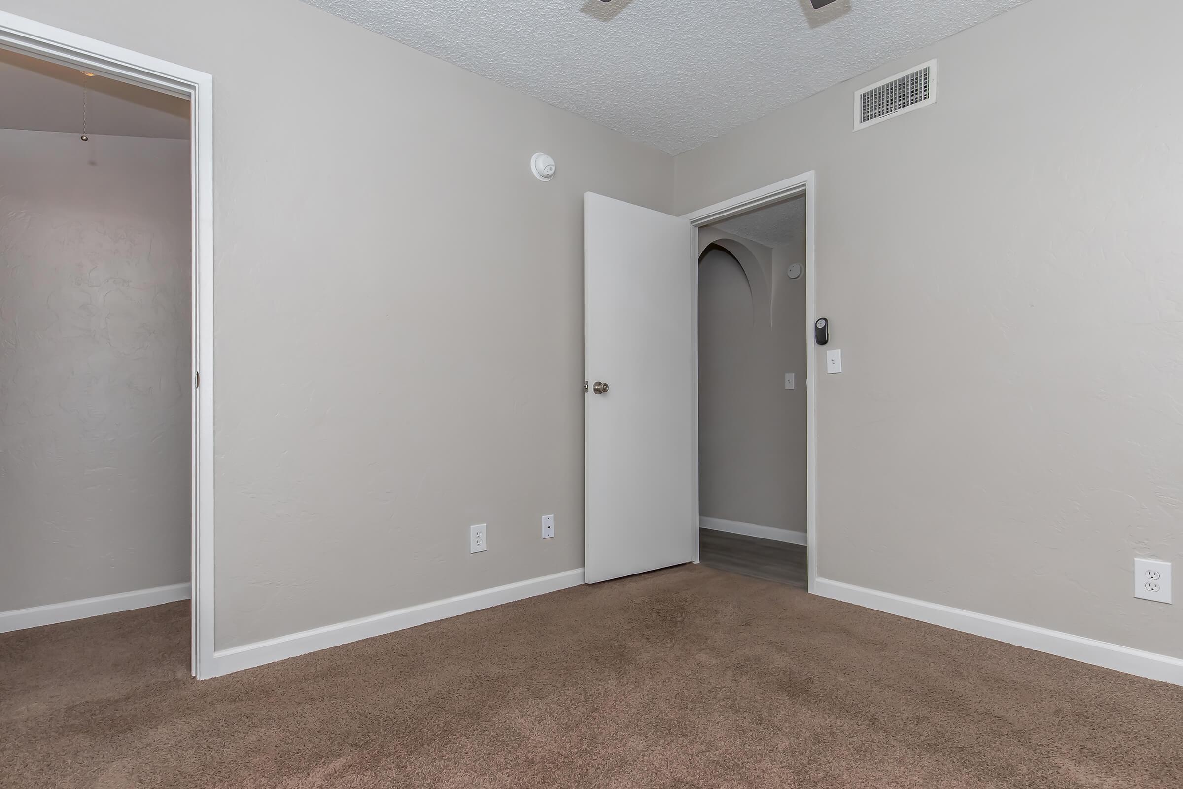 Interior view of a room featuring light gray walls and plush brown carpet. A white door is slightly ajar, leading to another area. There is a light smoke detector on the ceiling, and a small wall panel near the door. The room appears well-lit and clean, with minimal furnishings.