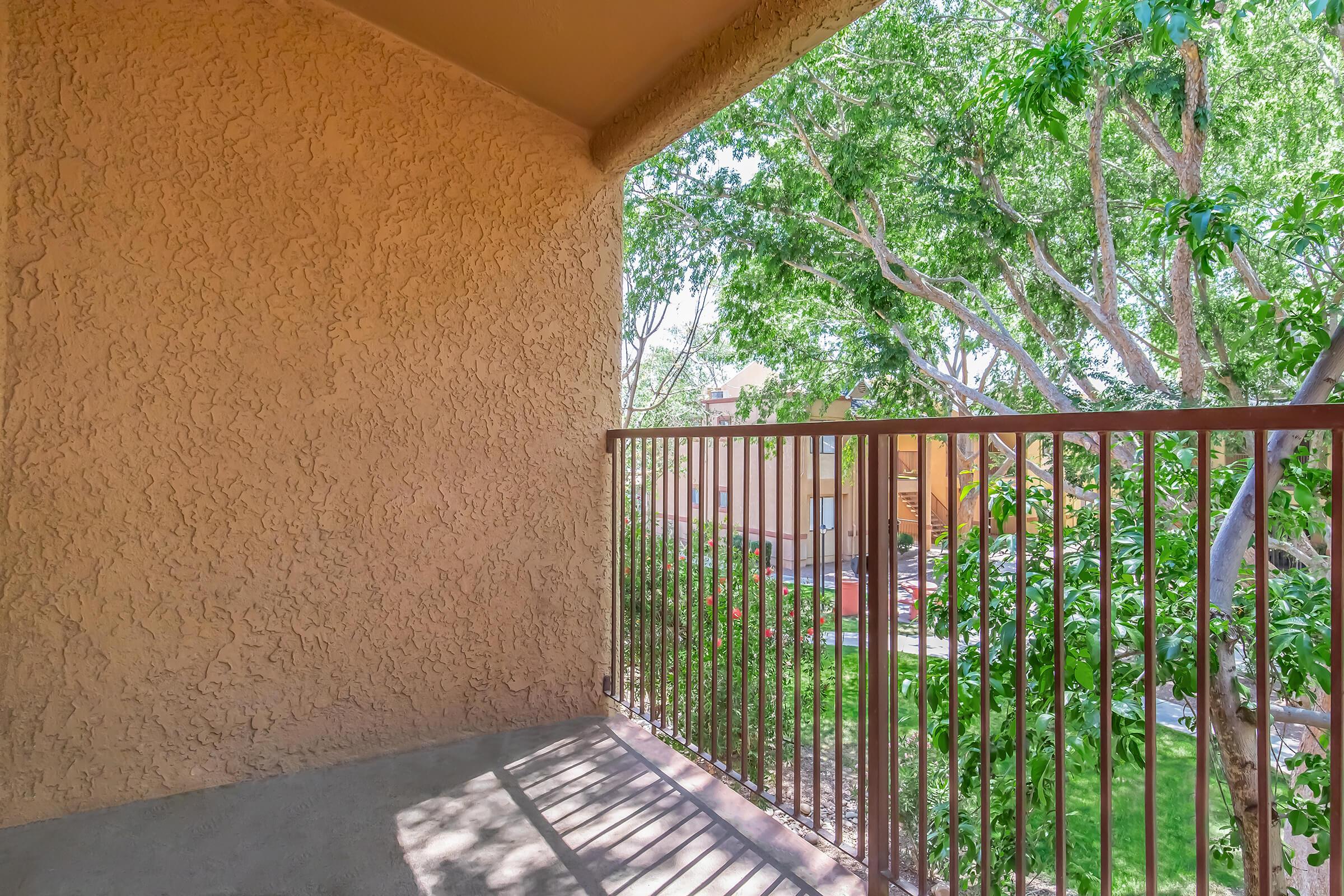 A balcony view featuring a textured wall and a metal railing overlooking lush greenery. Sunlight casts shadows on the concrete floor, while trees and foliage create a serene atmosphere in the background. In the distance, there are hints of nearby buildings.