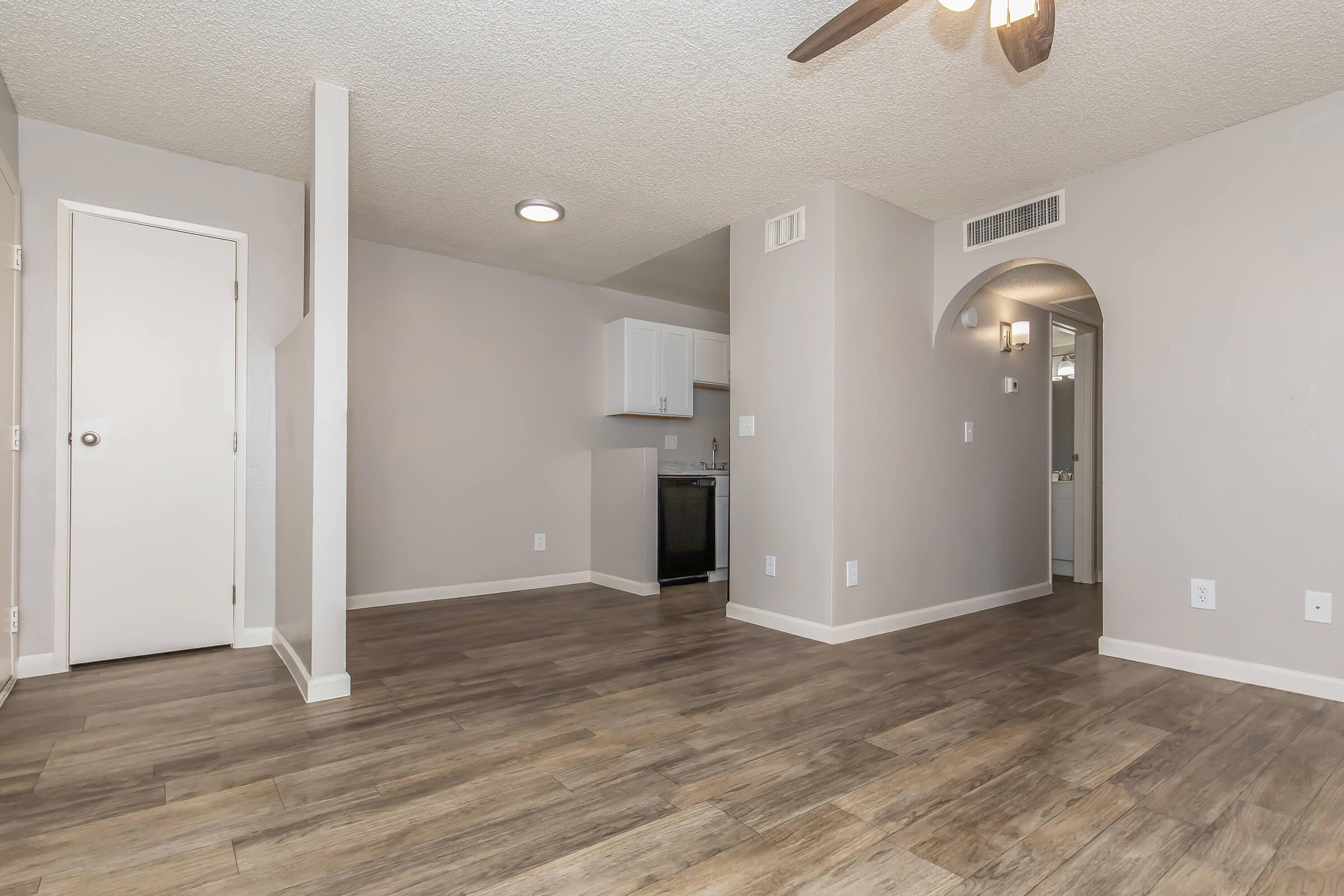 A spacious, modern living area featuring light-colored walls and wooden laminate flooring. The open layout includes a kitchen area with white cabinets, a ceiling fan, and doorways leading to other rooms. Natural light brightens the space, creating a welcoming atmosphere.