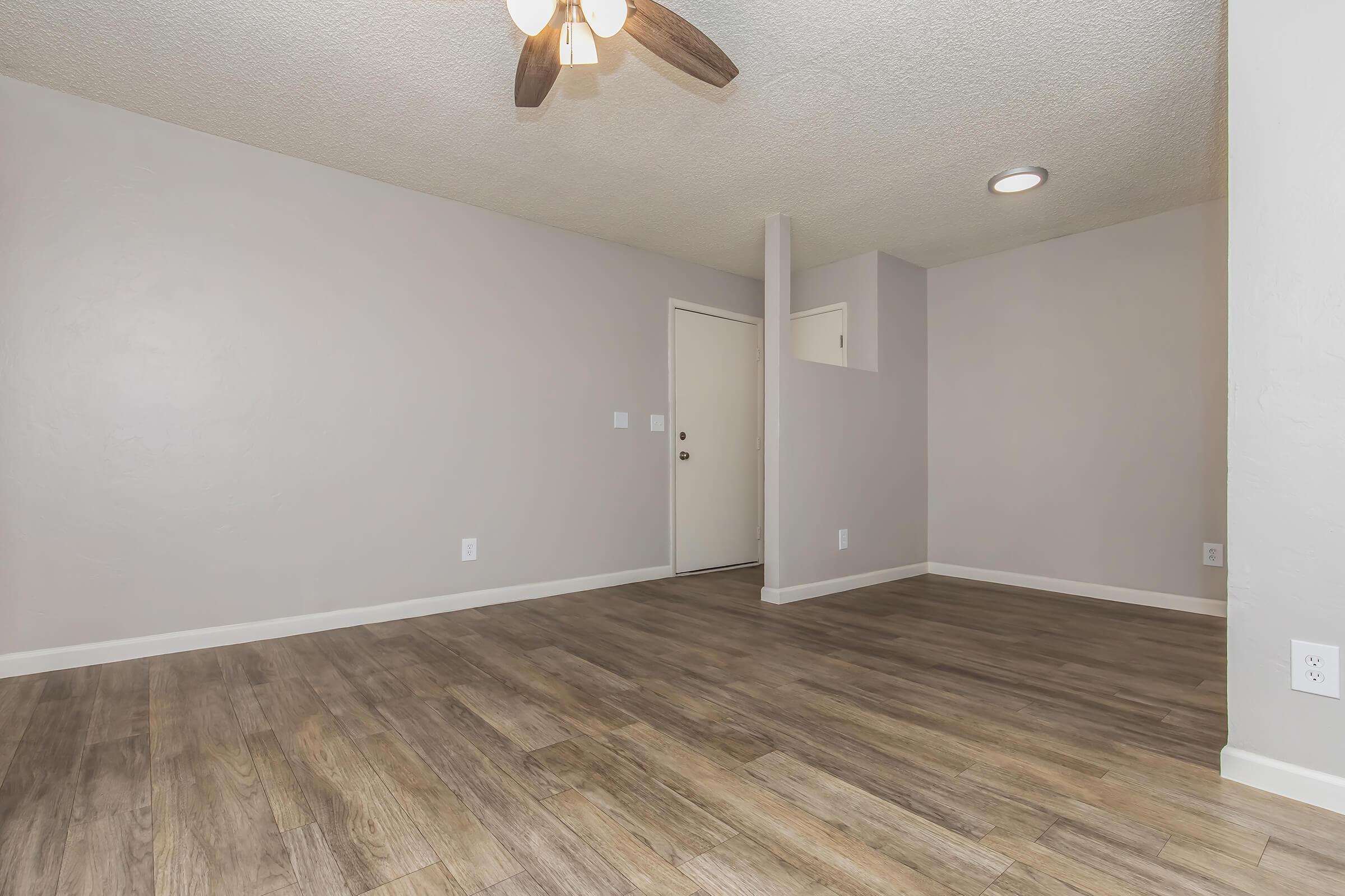 A spacious living area with light-colored walls and a wooden floor. There's a ceiling fan, a door leading outside, and a small wall nook. The room is well-lit with natural light, creating a welcoming atmosphere.