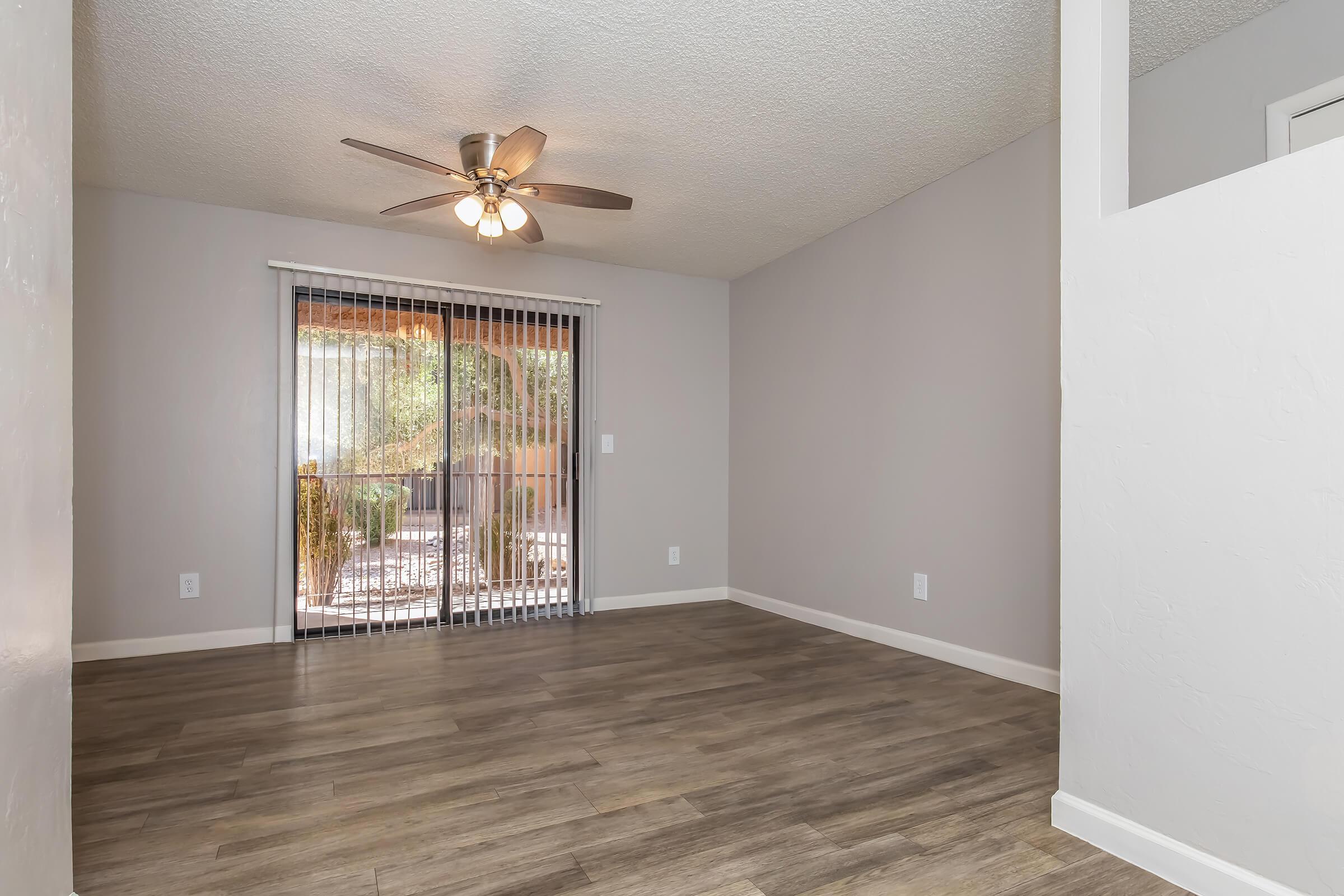 A spacious and well-lit living room featuring a ceiling fan, large sliding glass door with vertical blinds, and neutral-colored walls. The floor is made of light wood-like tiles. The room is empty, providing a blank canvas for potential furnishings and decor. A glimpse of an outdoor space is visible through the glass door.