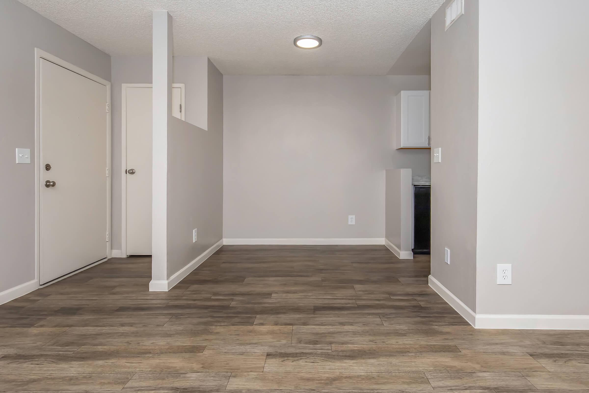 An empty interior space featuring a light gray color scheme. The floor is made of wood-like tiles, and there are two doorways visible. One door leads outside, while the other connects to a room. A small kitchen area with white cabinets is partially visible in the background. The overall atmosphere is bright and modern.