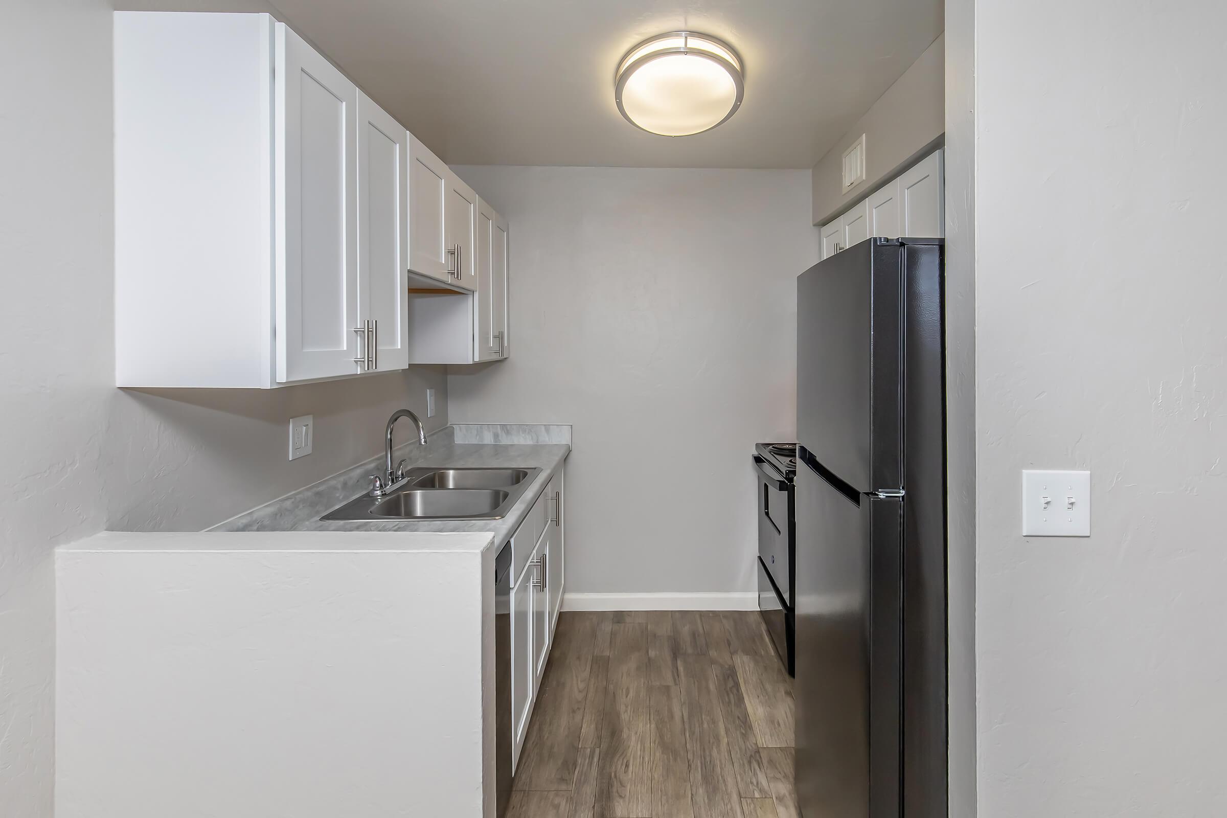 A modern kitchen featuring white cabinets, a stainless steel sink, and a black refrigerator. The walls are painted gray, and the flooring is wood-like. The space is illuminated by a ceiling light, and there is a stove visible next to the refrigerator.
