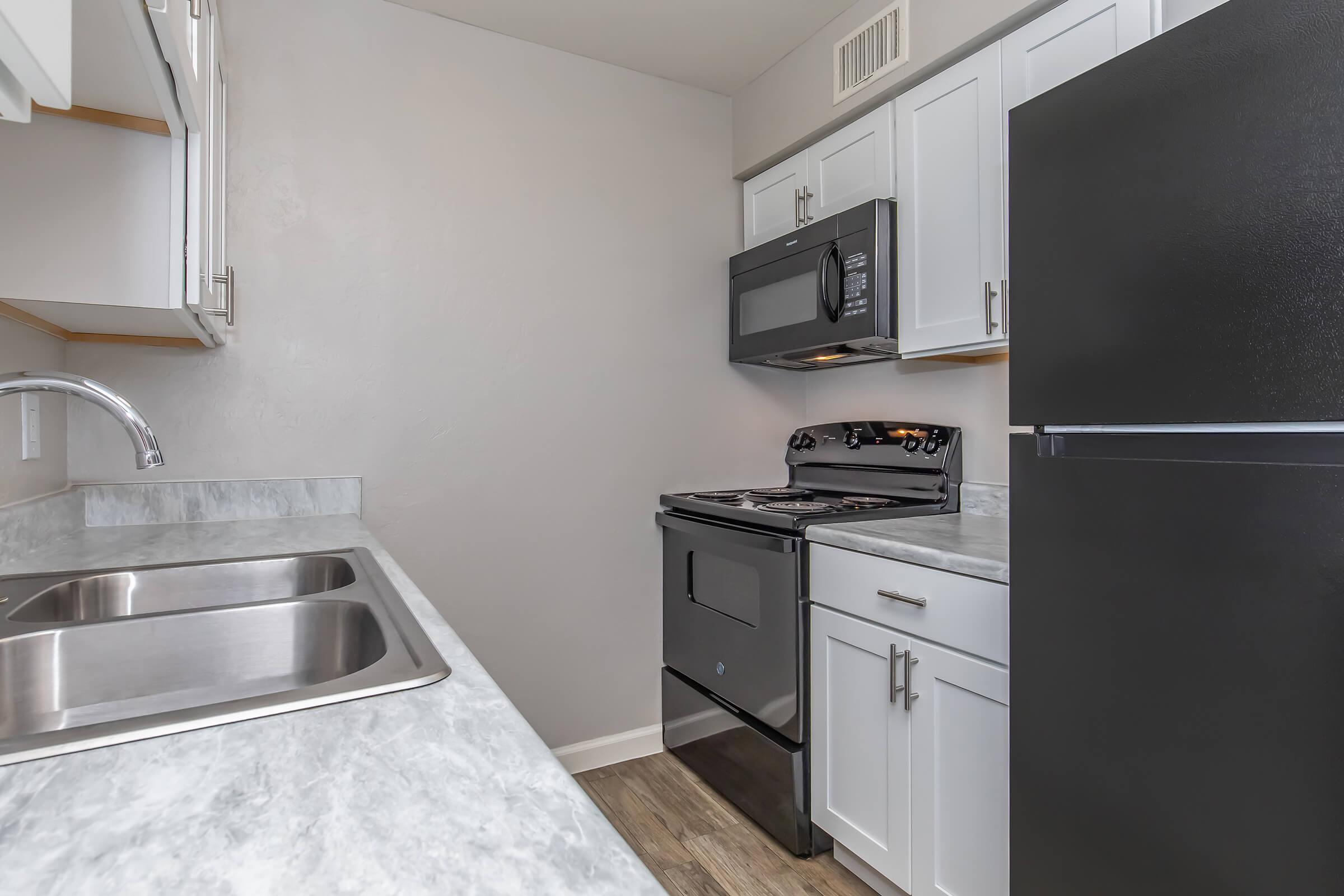A kitchen featuring white cabinets, a stainless steel double sink, a black stove with an oven, a microwave above it, and a black refrigerator. The countertop has a marble-like finish, and the walls are painted light gray. Natural light enhances the clean and modern look of the space.