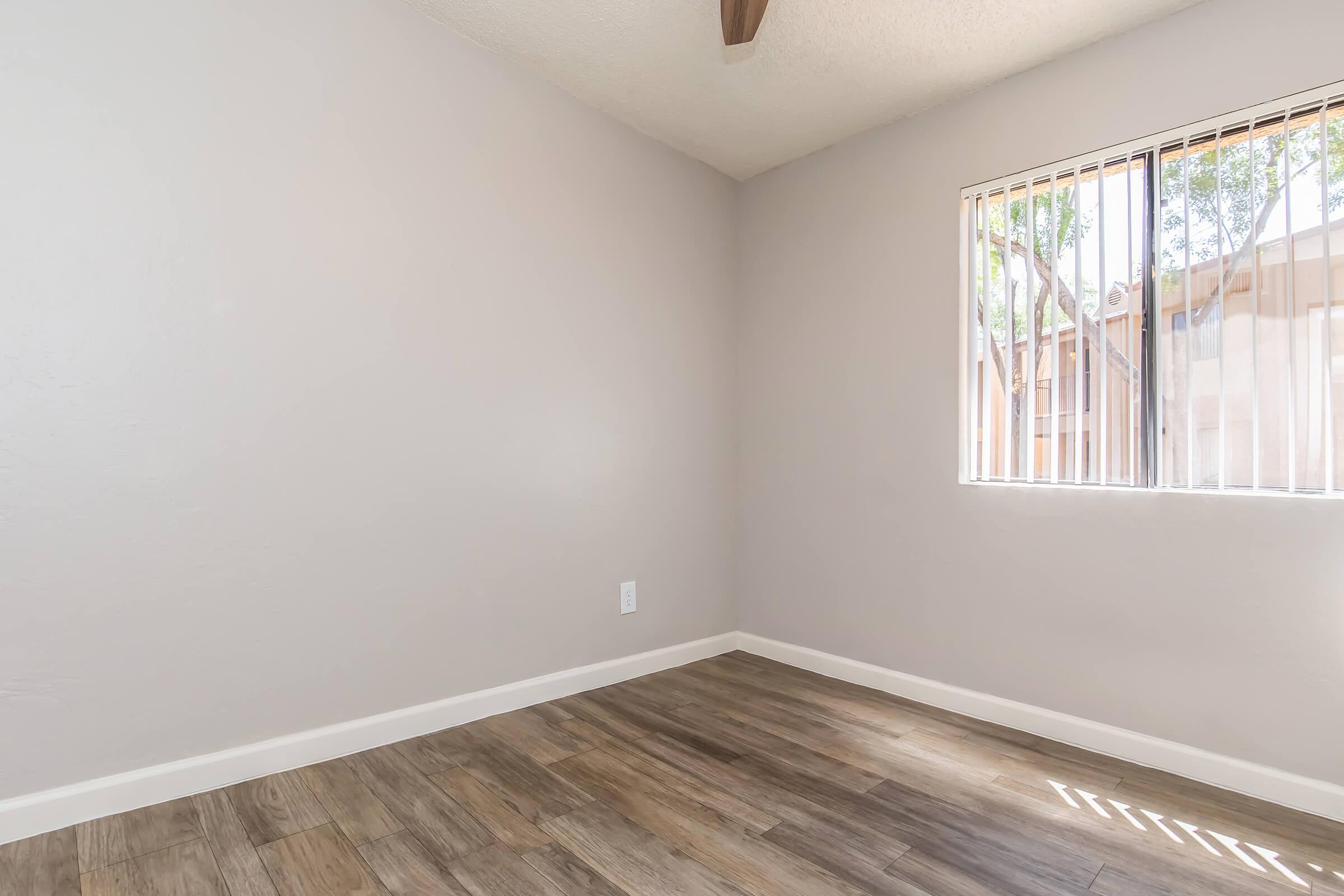 A vacant room with light gray walls and a wooden floor. There is a window on the right side with vertical blinds, allowing natural light to enter. The ceiling features a wooden beam, adding some architectural detail to the otherwise simple and clean space.