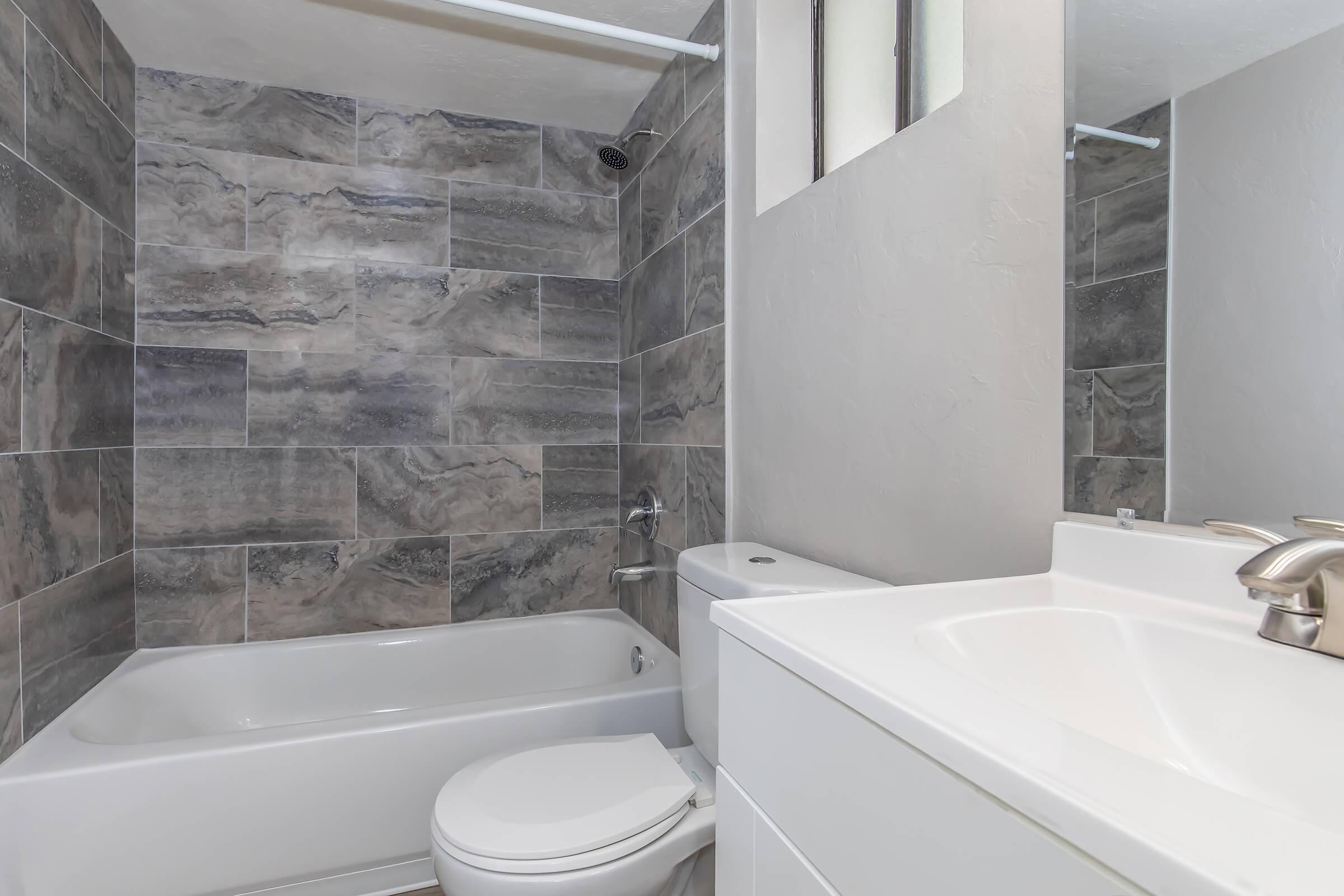 A modern bathroom featuring a shower and bathtub combination with gray stone tiles on the walls, a white toilet, and a sleek white sink with a faucet. The space is well-lit, showcasing a clean and contemporary design.