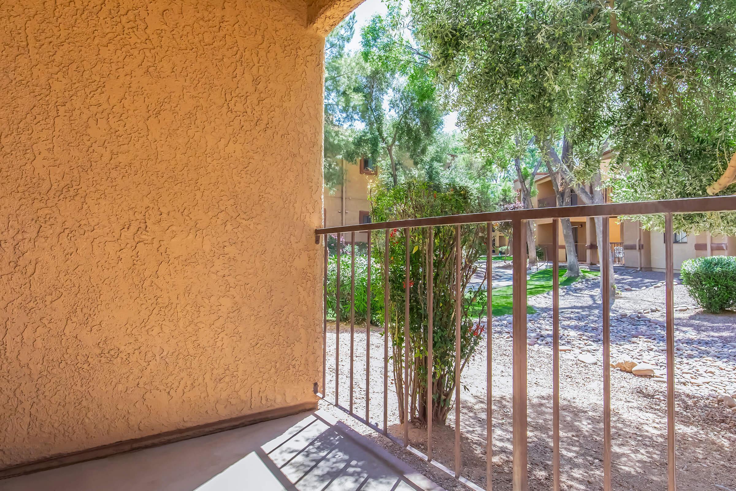 View from a balcony featuring a textured beige wall and a metal railing. Below, a landscaped garden is visible with shrubs and trees, creating a serene outdoor space. Soft sunlight filters through the leaves, casting gentle shadows on the ground. The environment looks well-maintained and inviting.