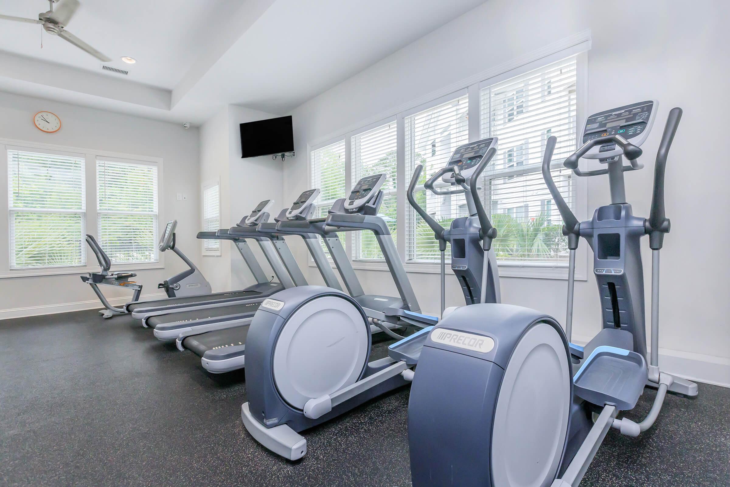 A modern gym area featuring several elliptical machines and stationary bicycles lined up against large windows. The walls are painted in light colors, and there is a clock and a television mounted for entertainment. The flooring is dark rubber, providing a functional workout space.