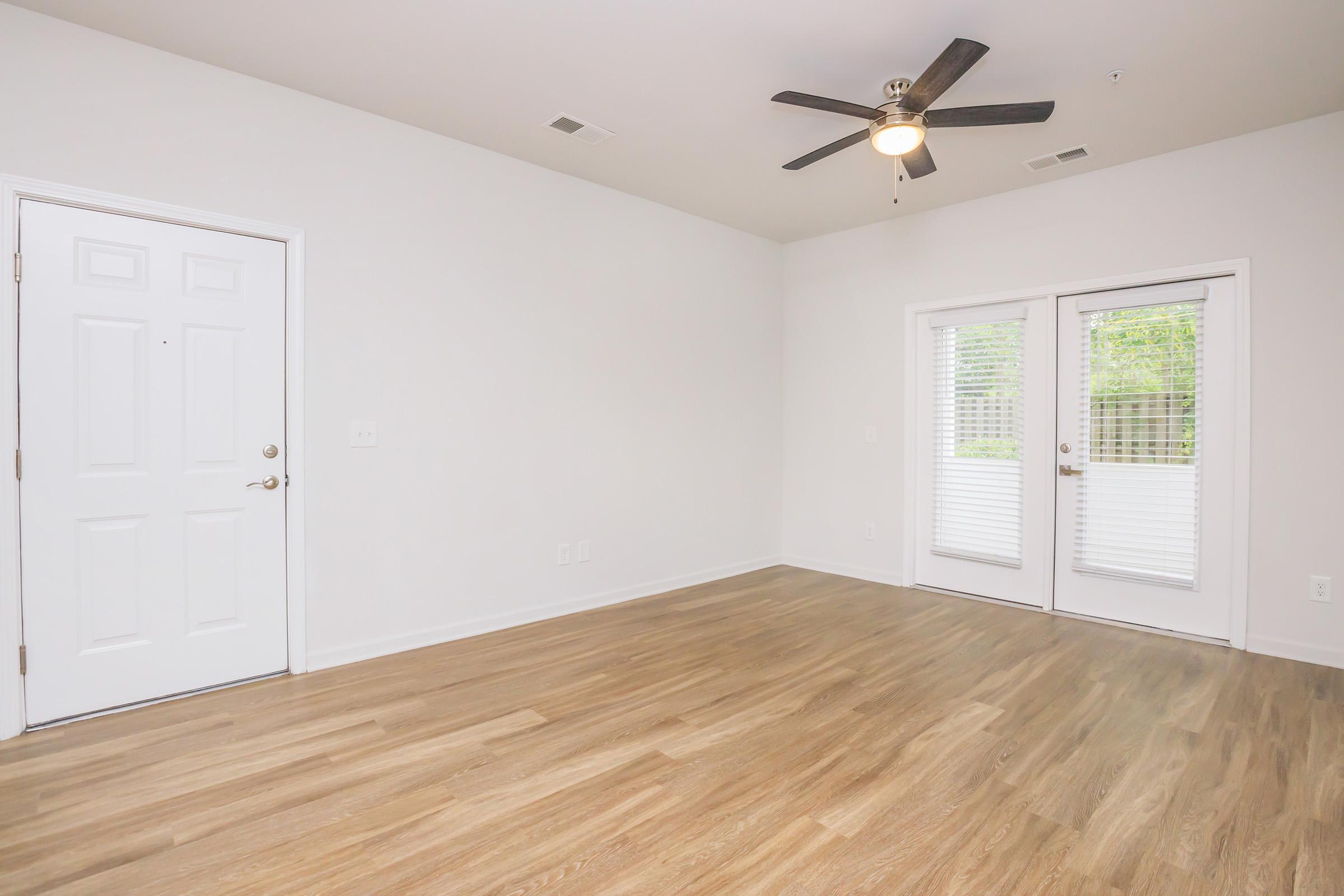 A bright, empty room featuring light wood flooring, a ceiling fan, and a front door on one side. There are sliding glass doors on the opposite side, leading to a small outdoor area with greenery visible outside. The walls are painted white, creating a clean, spacious feel.