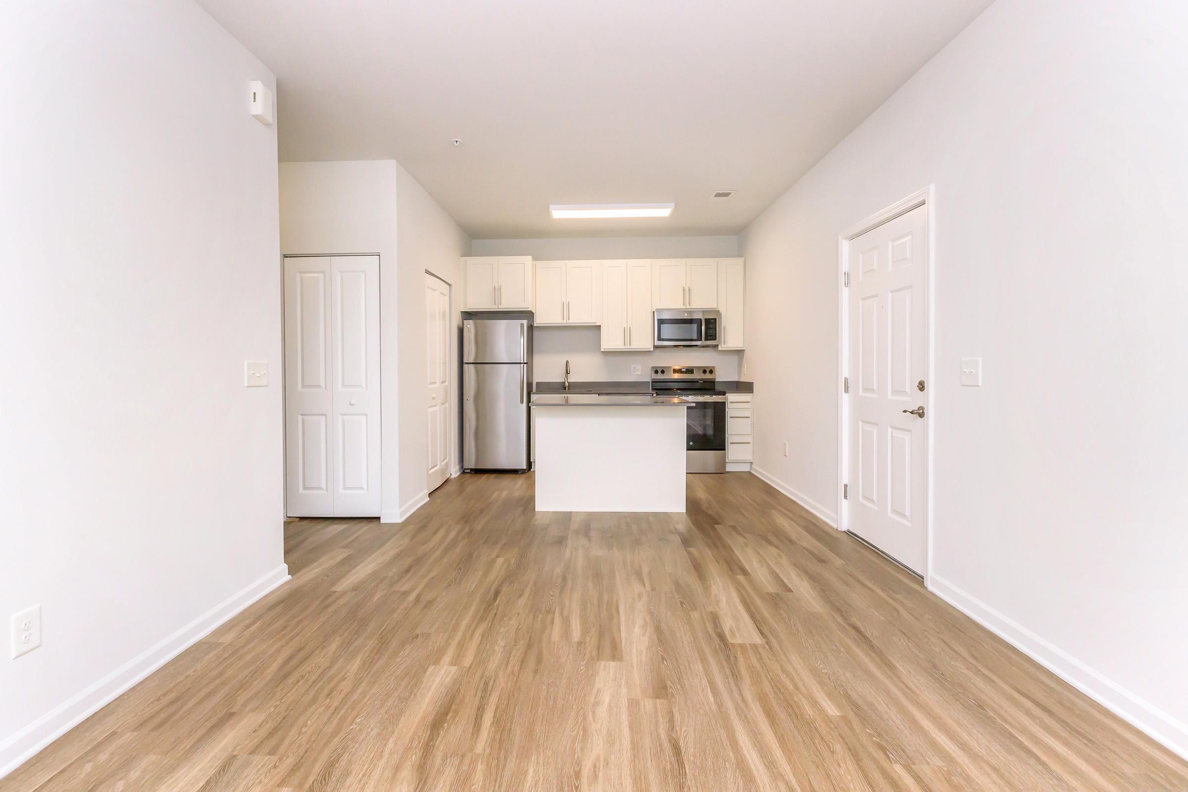 A modern, open-concept kitchen and living area featuring light-colored walls, wooden flooring, and a centrally placed island. The kitchen includes stainless steel appliances, white cabinetry, and a door leading to a closet. Natural light brightens the space, creating an inviting atmosphere.