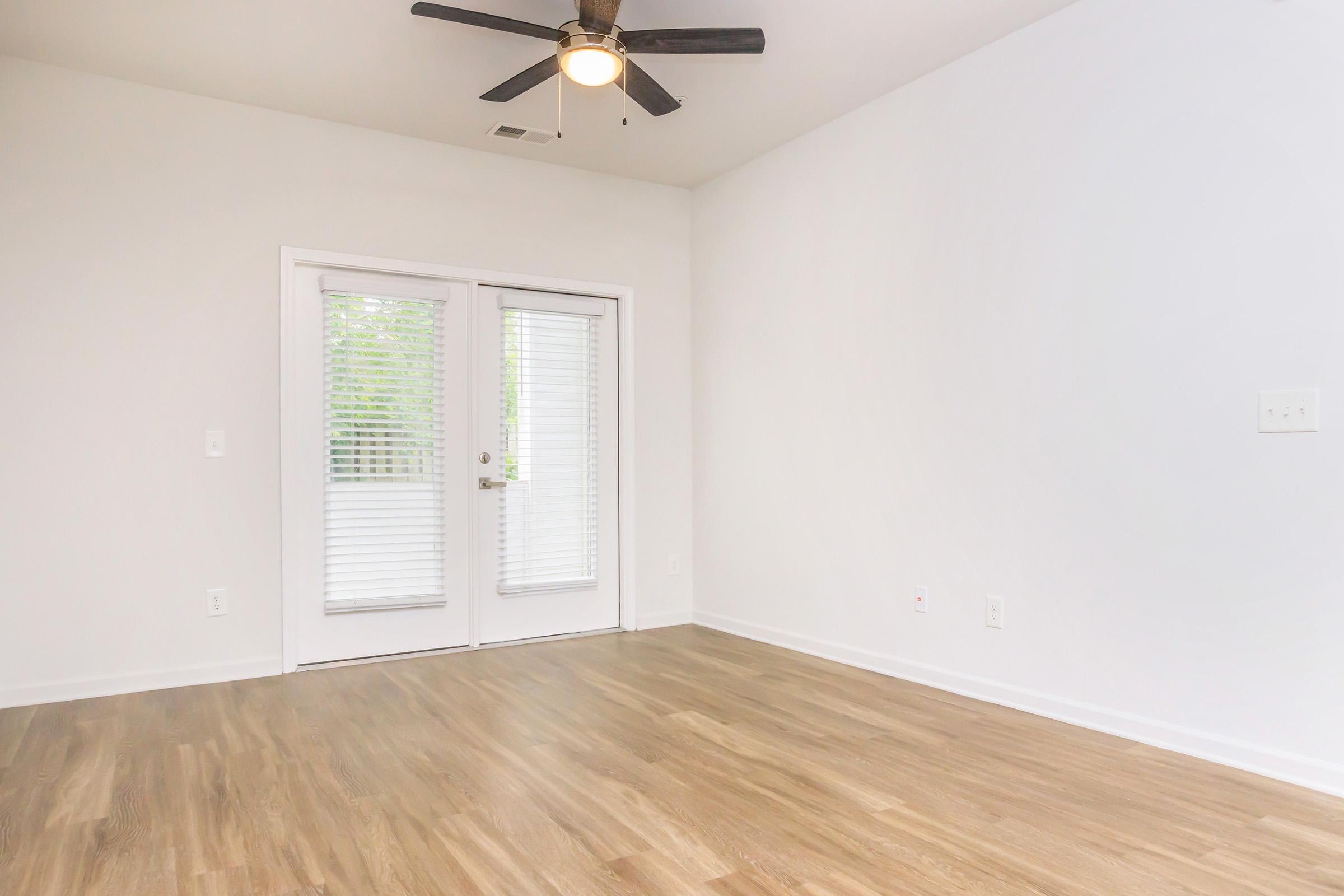 A spacious, empty room with light-colored walls and wooden flooring. A ceiling fan is installed above, and two sets of glass doors with blinds are visible, leading to an outdoor area. Natural light brightens the room, enhancing its airy feel.