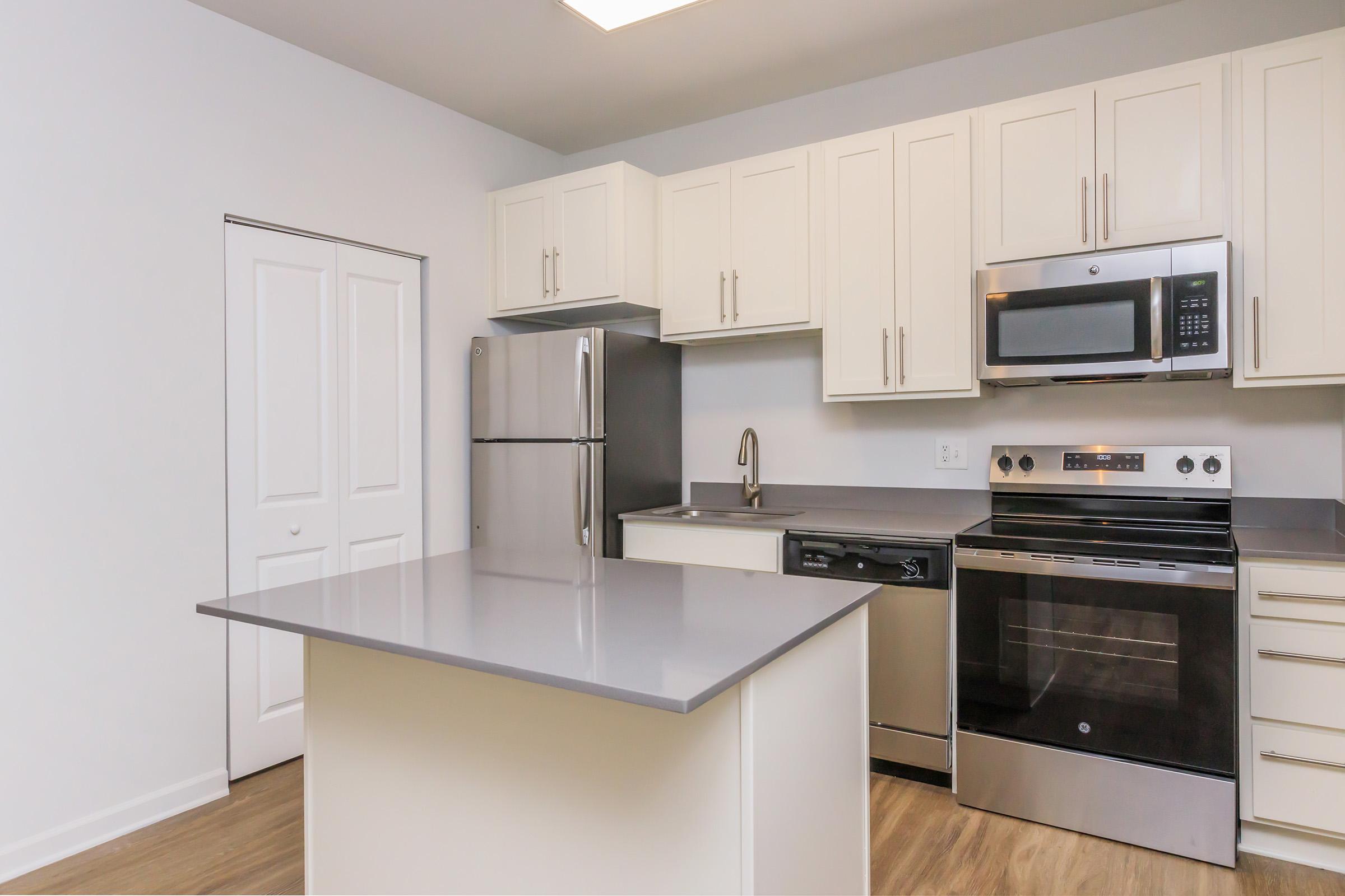 Modern kitchen featuring white cabinets, stainless steel appliances including a refrigerator, microwave, and oven. A gray countertop island is in the center, with a sink and dishwasher nearby. The walls are painted light gray, and there is a closet door visible to the left. The flooring is light wood.