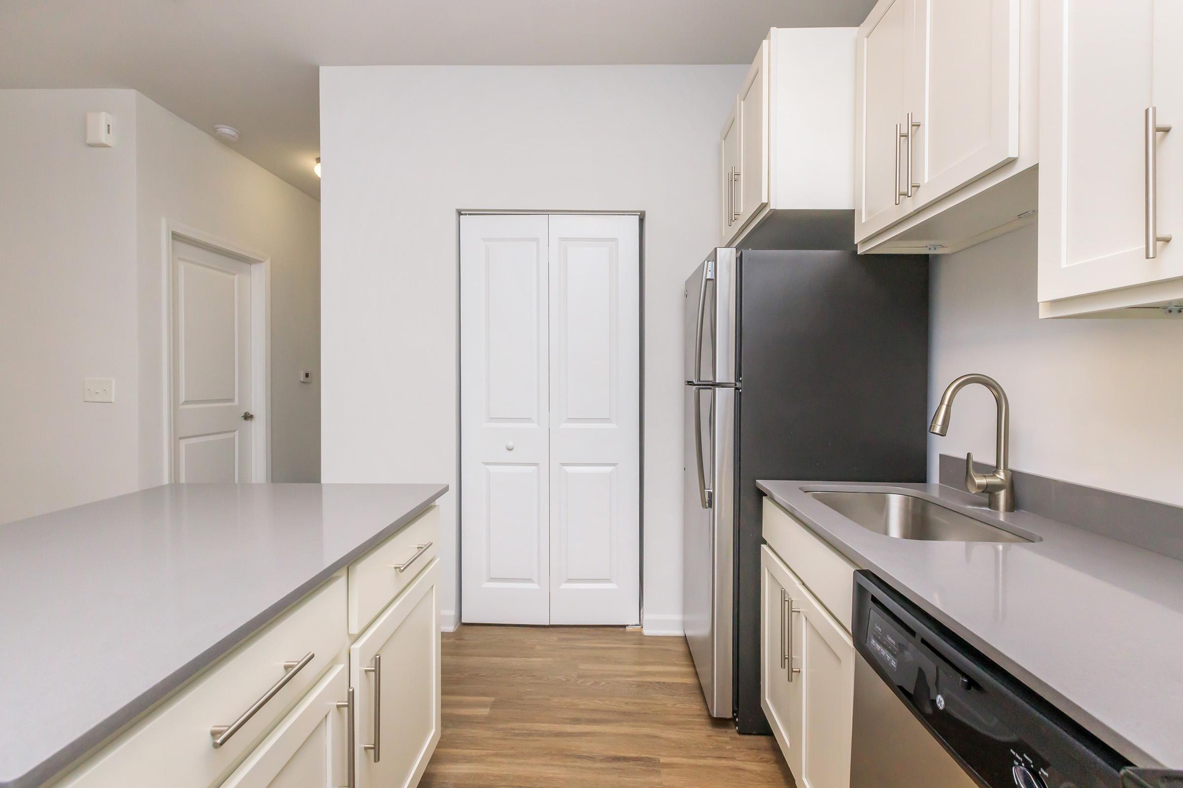 A modern kitchen featuring white cabinetry, stainless steel appliances, a gray countertop, and a single-basin sink. The space is well-lit, with a door leading to a closet visible in the background. The flooring is a warm wood-like design, adding to the contemporary aesthetic.