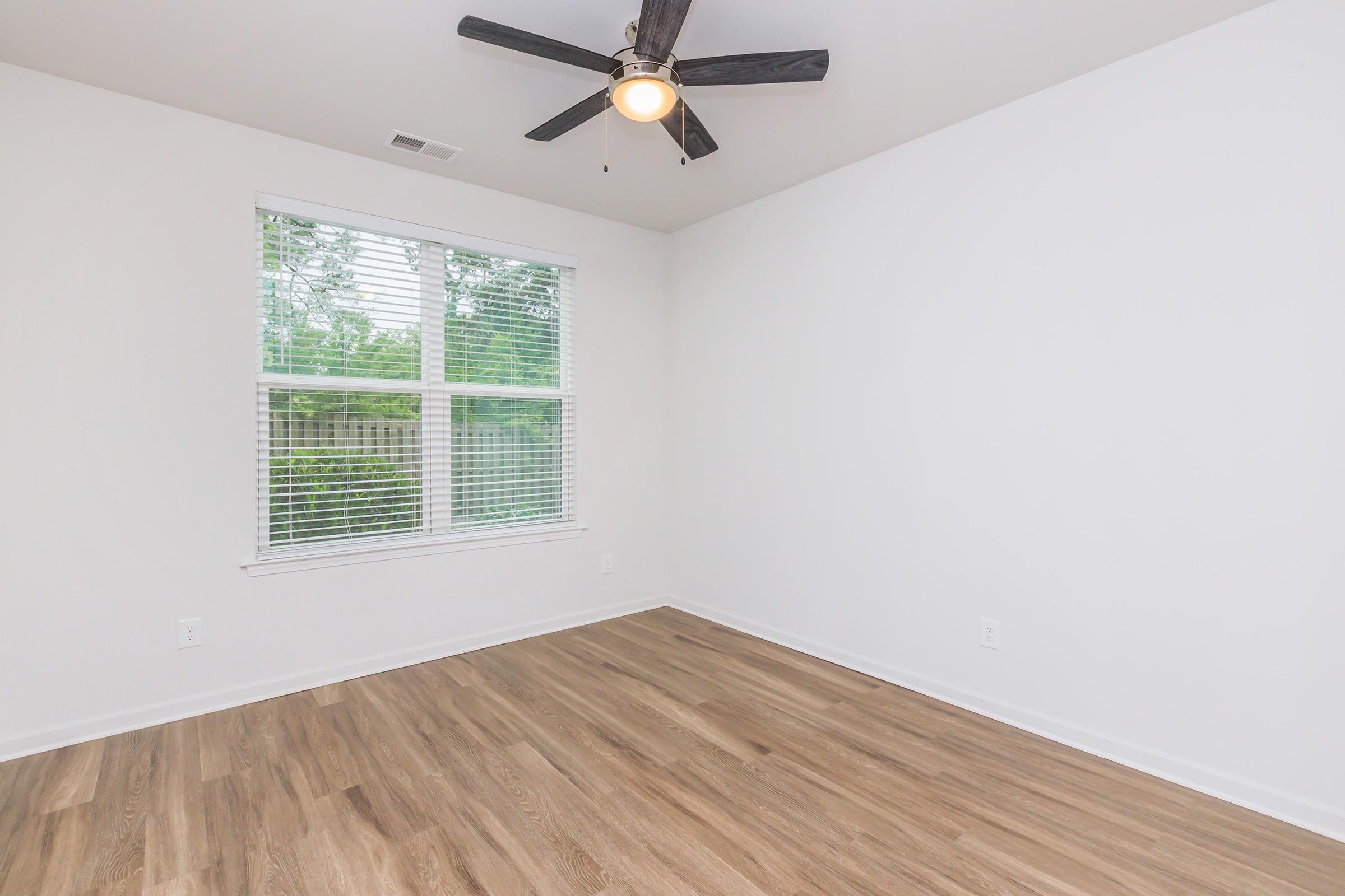 A spacious, empty room with light hardwood flooring, a ceiling fan, and a large window featuring white blinds, allowing natural light to illuminate the space. The walls are painted white, creating a bright and airy atmosphere, suitable for various uses such as a bedroom or office.