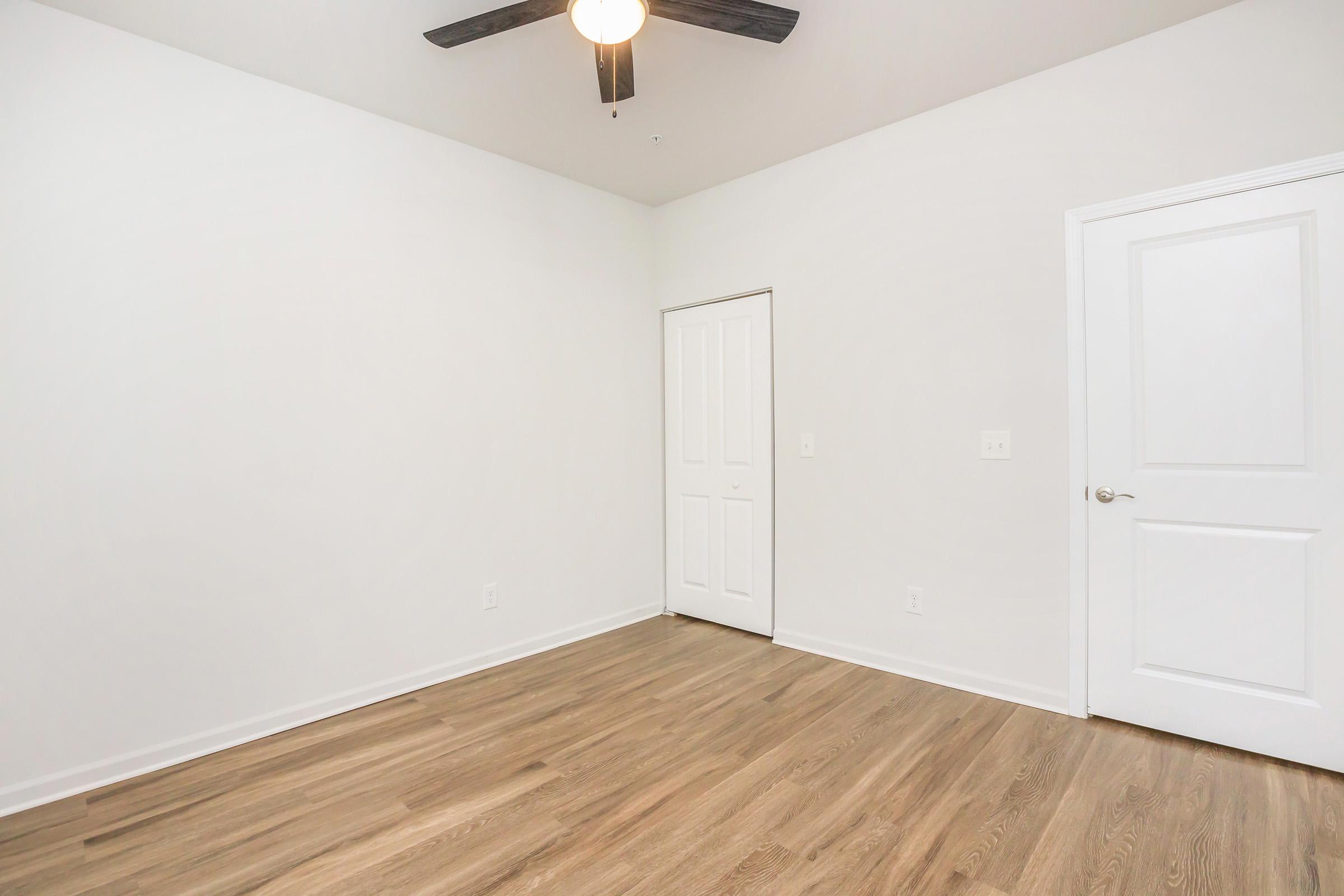 A bright, empty room with white walls and a ceiling fan. The floor is made of light-colored wood. A closed door is visible on the left side of the image. The overall atmosphere is clean and minimalistic, ideal for personal decoration or furnishing.