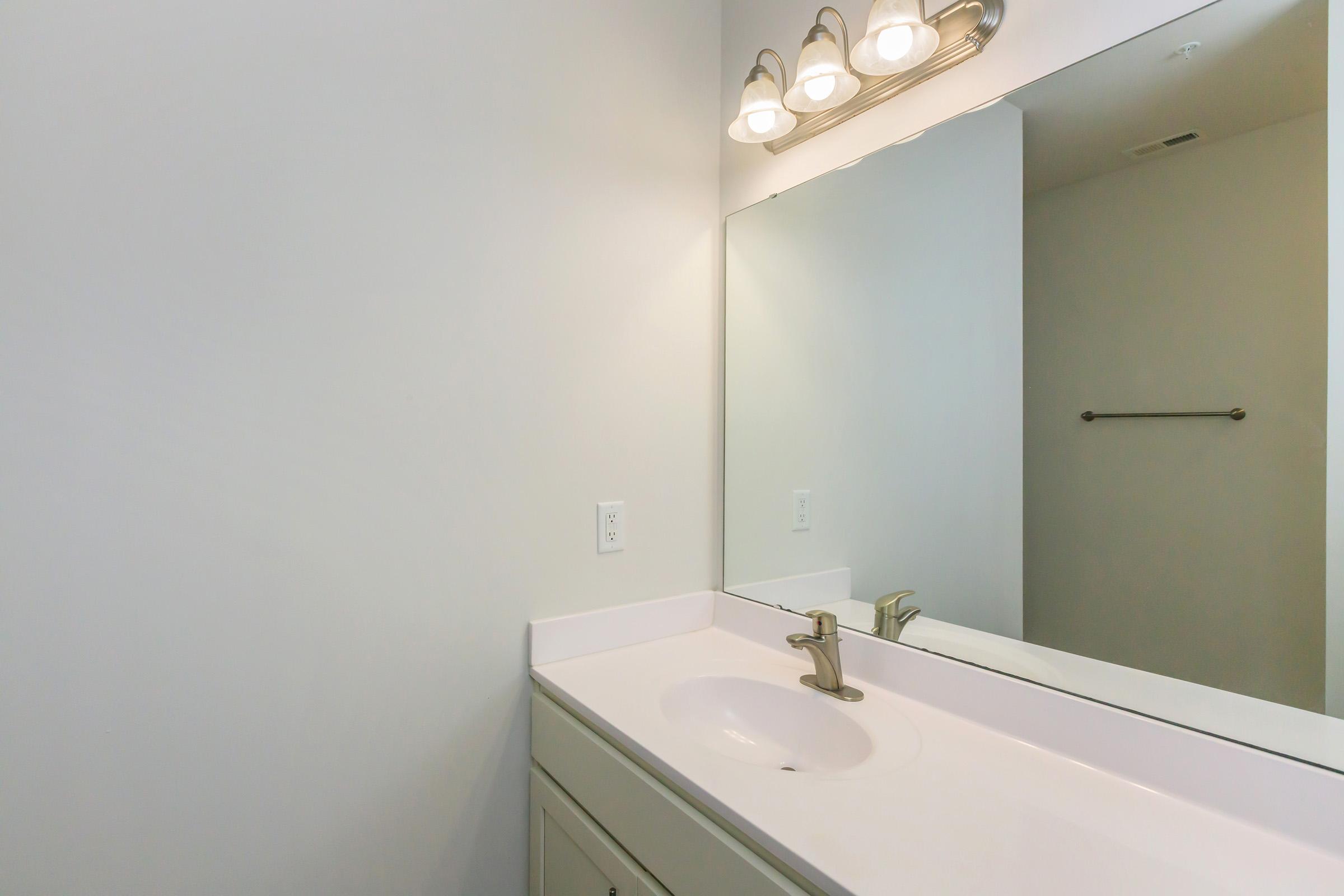 A clean bathroom vanity featuring a white sink and a large mirror above it. Three light fixtures are mounted above the mirror. The walls are painted a light color, and there is a towel bar visible on the right side of the image. The overall aesthetic is simple and modern.