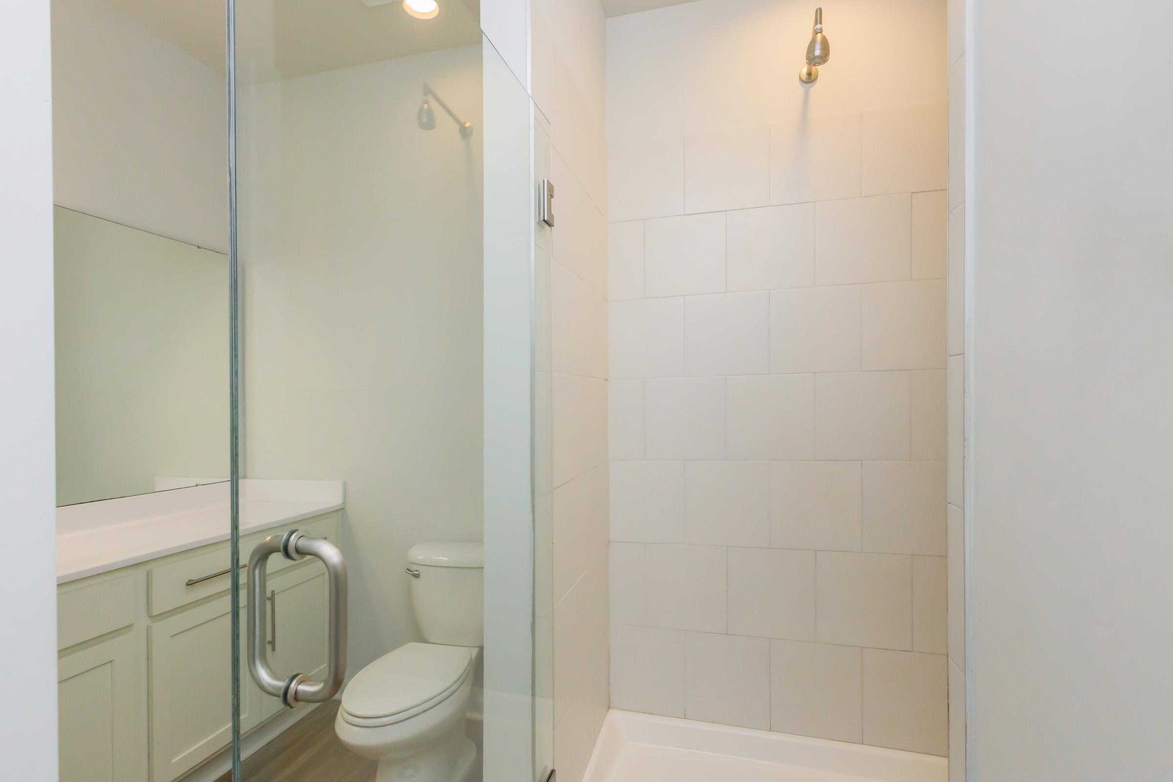A modern bathroom featuring a glass-enclosed shower with white tile, a wall-mounted light fixture, and a sink with cabinetry. A toilet is visible beside the shower, and the overall design is sleek and minimalistic. Natural light enhances the spacious feel of the area.