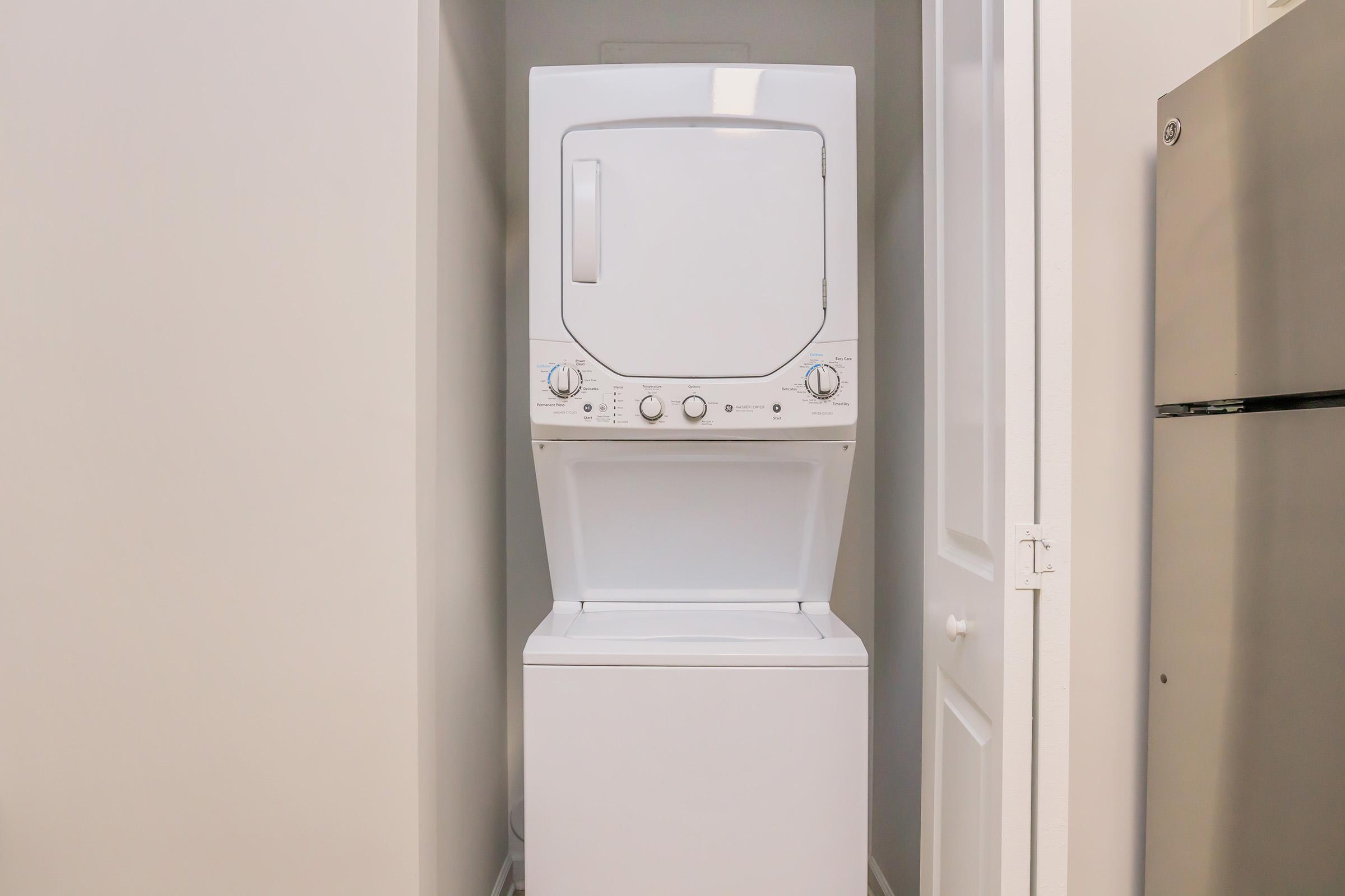 A white stacked washer and dryer located in a narrow laundry nook, with light-colored walls surrounding it. The door to the nook is open, and a silver refrigerator is visible to the right.