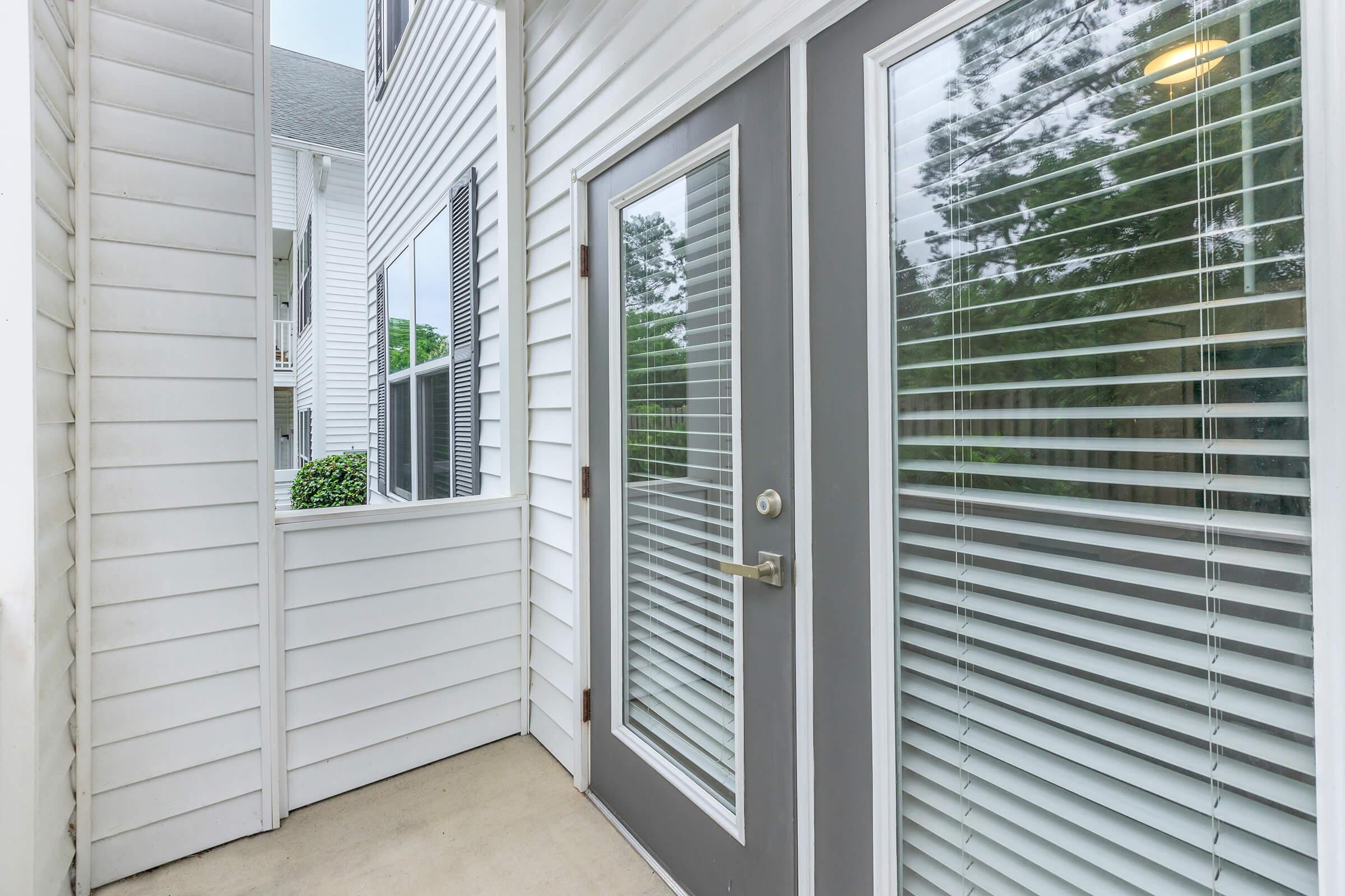 A modern entryway featuring a glass door with horizontal blinds, framed by white siding. The entrance is well-lit, with a small porch area visible, and greenery is seen through the glass.