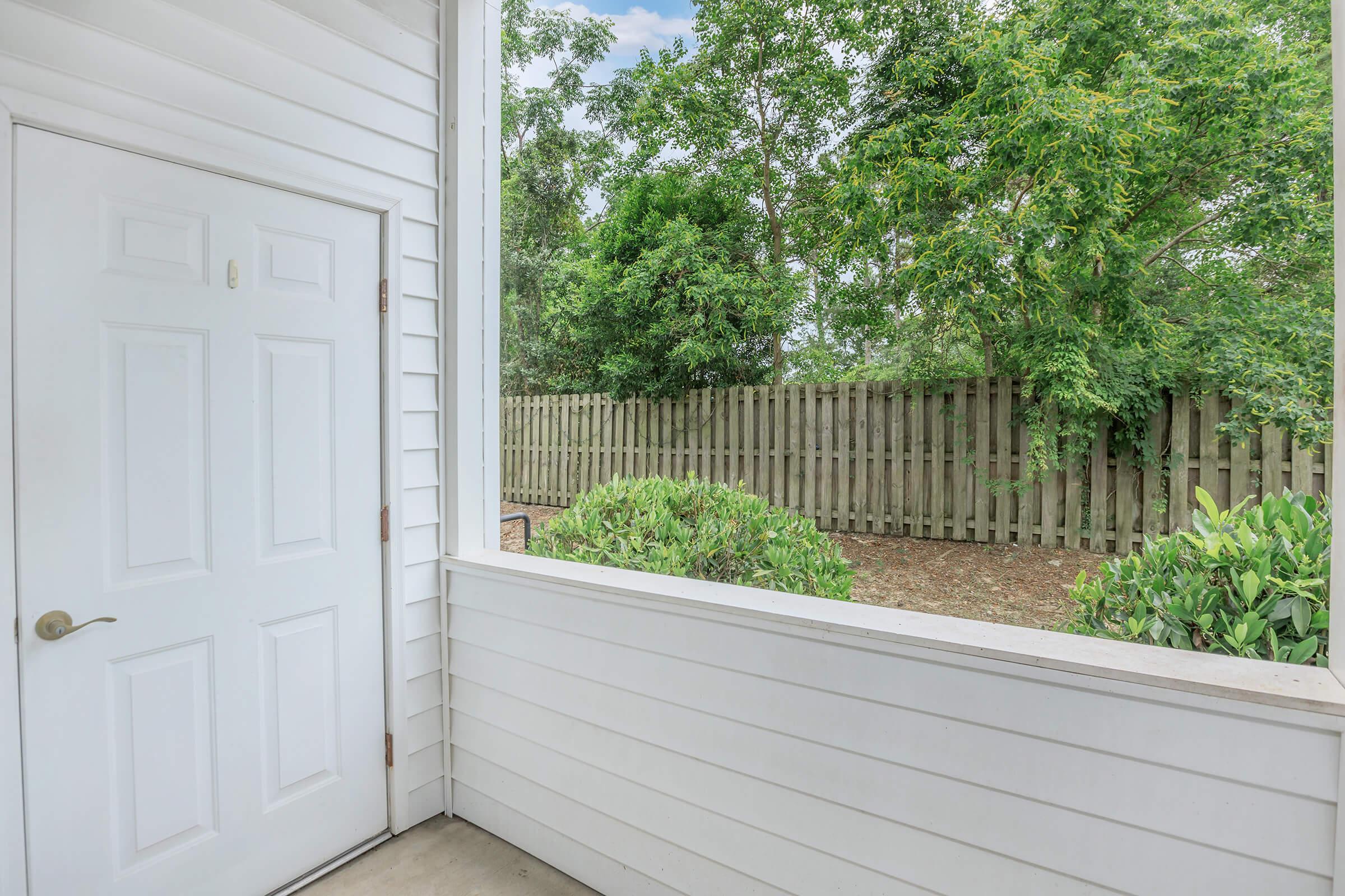 View of a small outdoor area from a porch, featuring a white door and a fenced yard with lush green trees and bushes. The scene is bright and serene, highlighting the natural surroundings with a clear sky and well-maintained landscaping.