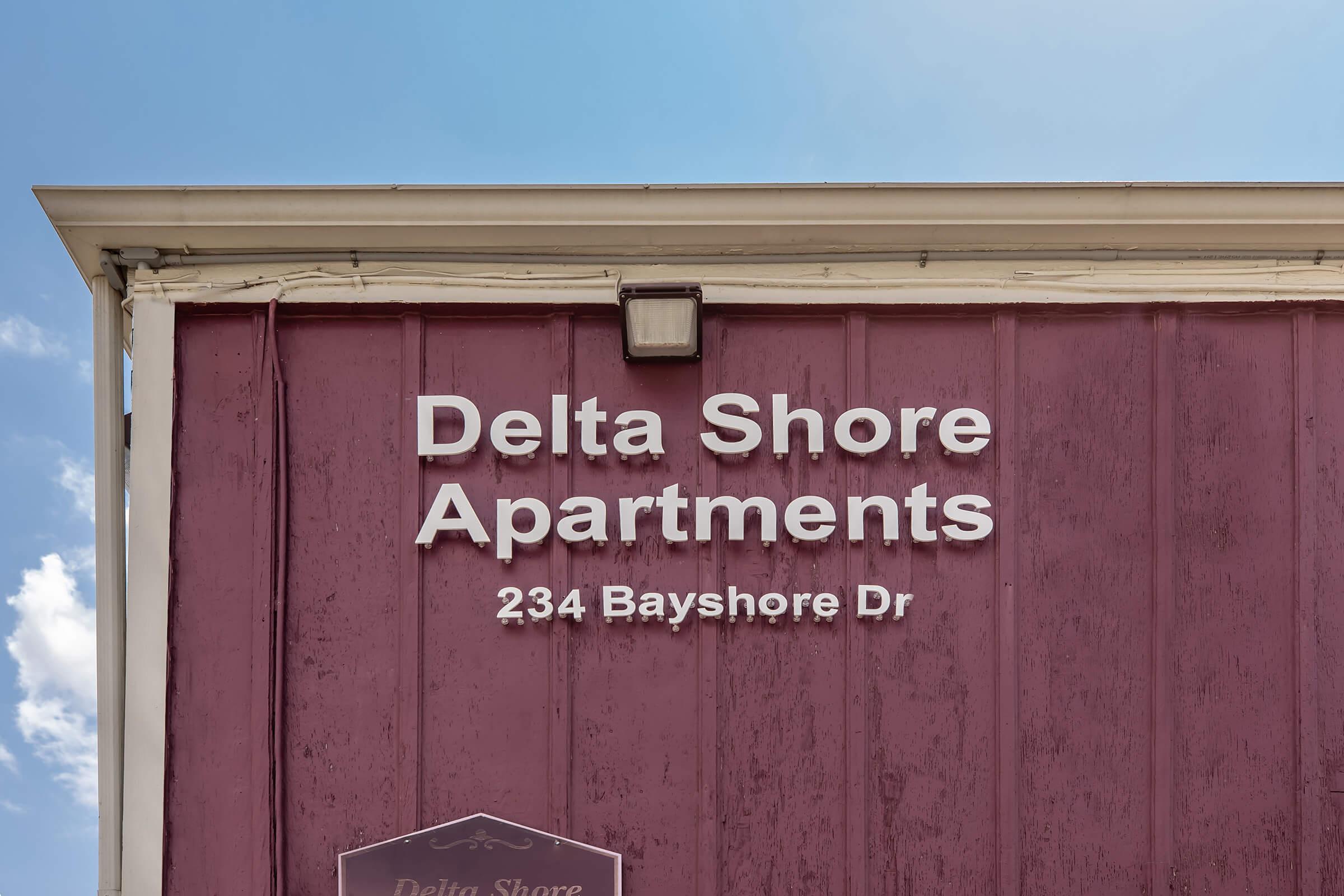 Signage for Delta Shore Apartments located at 234 Bayshore Drive. The building features a purple exterior and clear, white letters displaying the apartment name and address against a blue sky.
