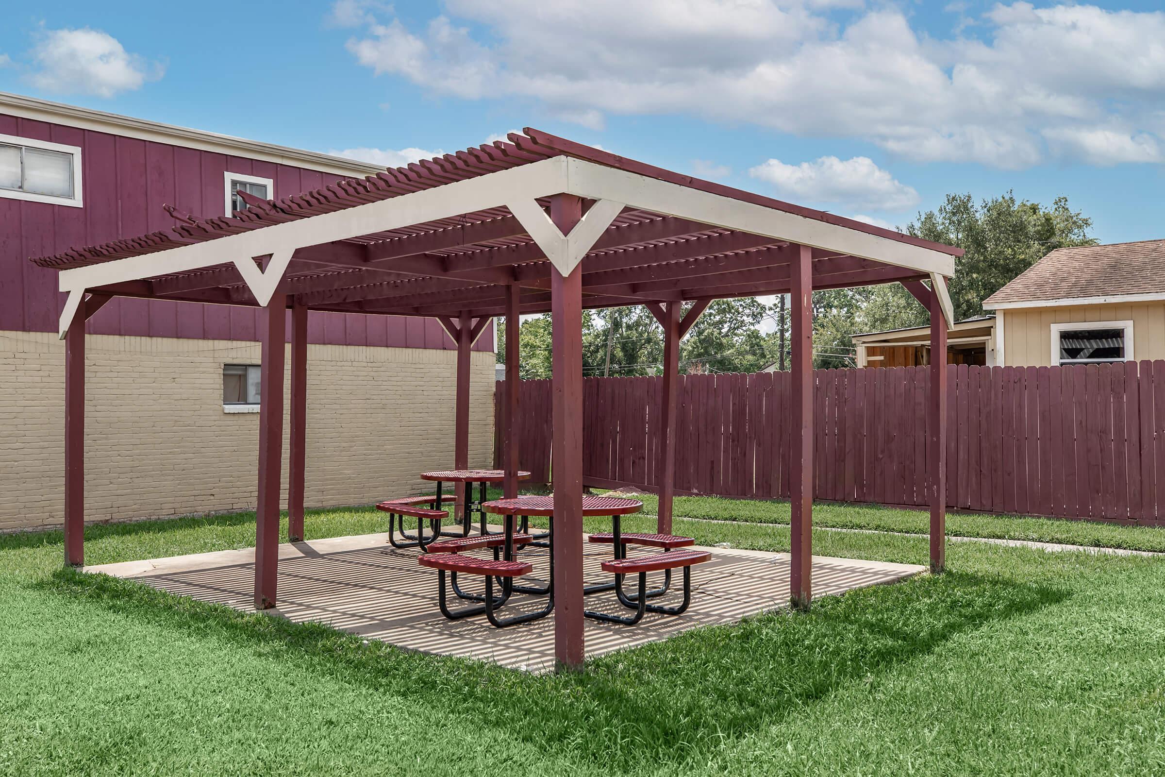 A shaded picnic area featuring a wooden pergola with several red picnic tables. The structure is surrounded by green grass and nearby buildings, with a wooden fence in the background. The sky is partly cloudy, adding to the outdoor ambiance.