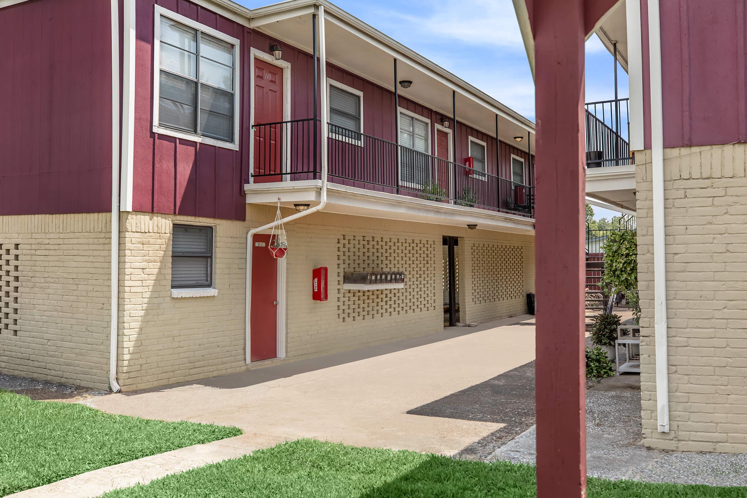 A view of an apartment building featuring red and beige exteriors. The ground level has a concrete pathway with grass on the sides, while several doors lead to individual units, and a row of mailboxes is visible on the wall. The scene is bright and well-lit, showcasing a clean and inviting atmosphere.