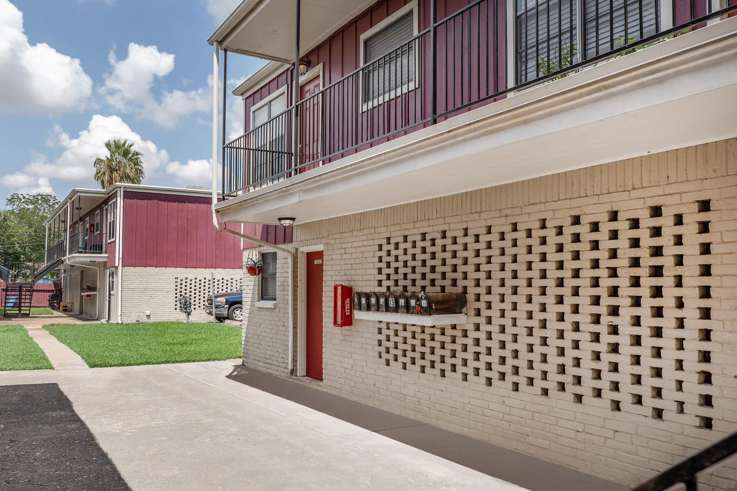 A view of a residential building with a two-story structure featuring a balcony above and a textured brick wall below. A mailbox area is visible on the wall, and surrounding grassy space is neatly maintained. The sky is partly cloudy, adding to the outdoor atmosphere.