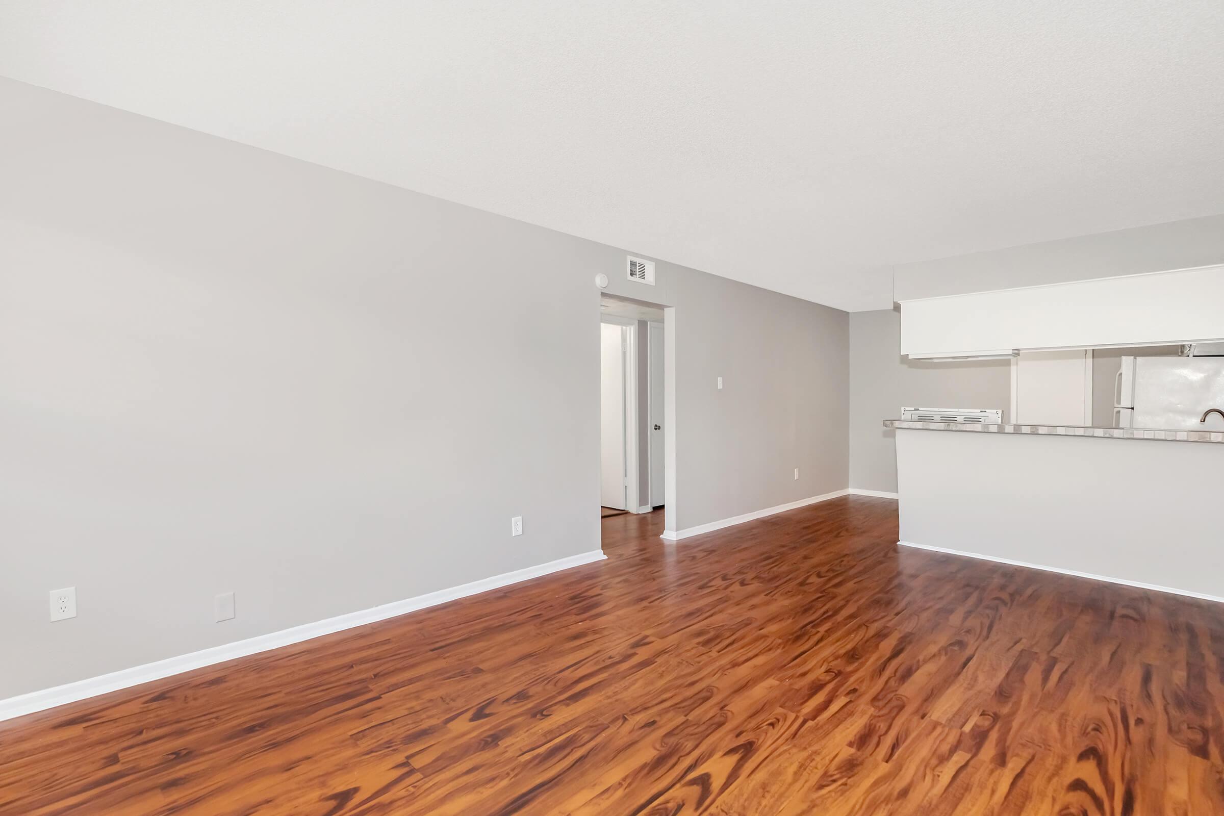 Spacious interior of an empty room featuring light gray walls and a shiny wooden floor. A doorway leads to another room, while an open kitchen area is visible in the background. The overall ambiance is bright and modern, making it suitable for various design styles.