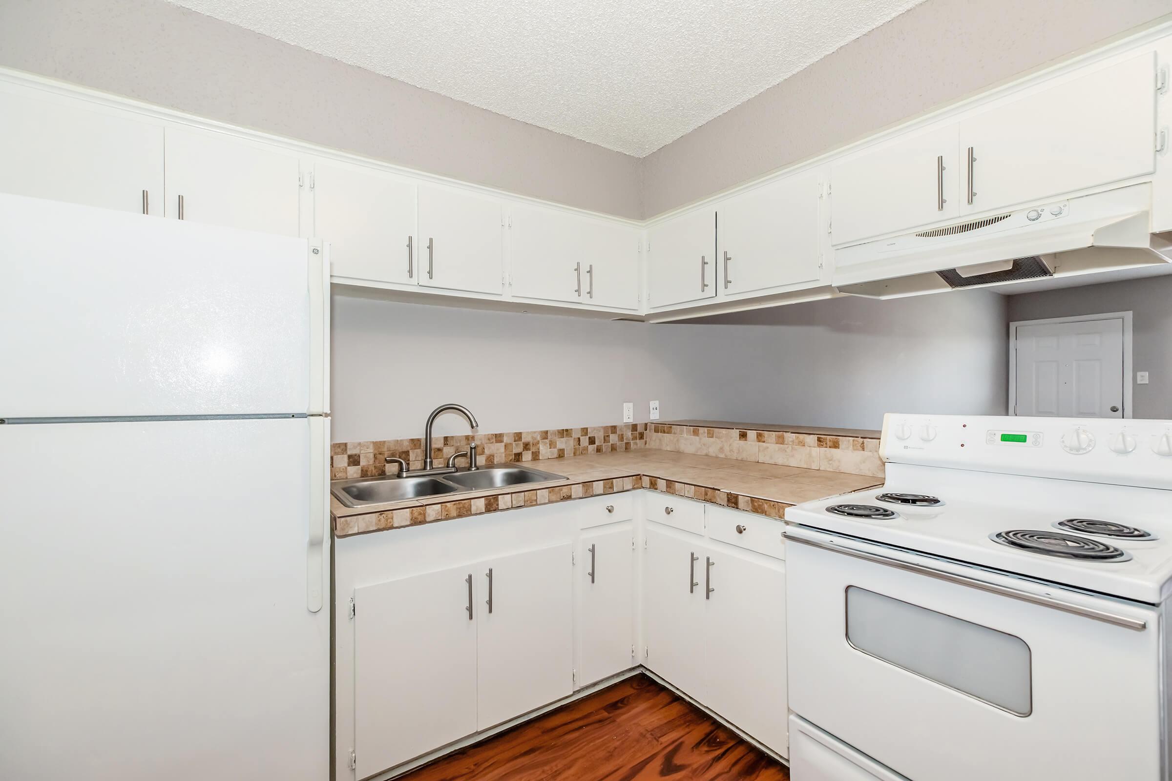 A well-lit kitchen featuring white cabinetry, a white refrigerator, a double sink with a chrome faucet, and a white stove. The countertops are beige with a decorative backsplash. The floor is wooden, and the room has a neutral-colored wall and an entrance visible in the background.