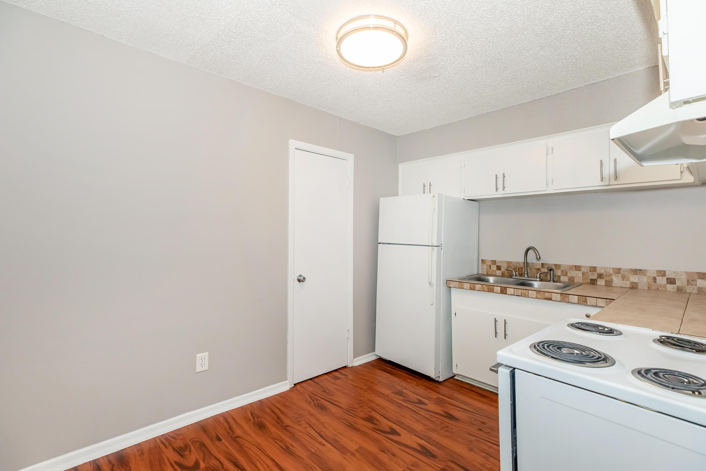 A simple kitchen featuring light gray walls, a white refrigerator, and a white stove with four burners. The countertop is tiled, and there is a stainless steel sink. The flooring is a warm, polished wood finish. A door is visible, leading to another room or area. Hanging light fixture above provides illumination.