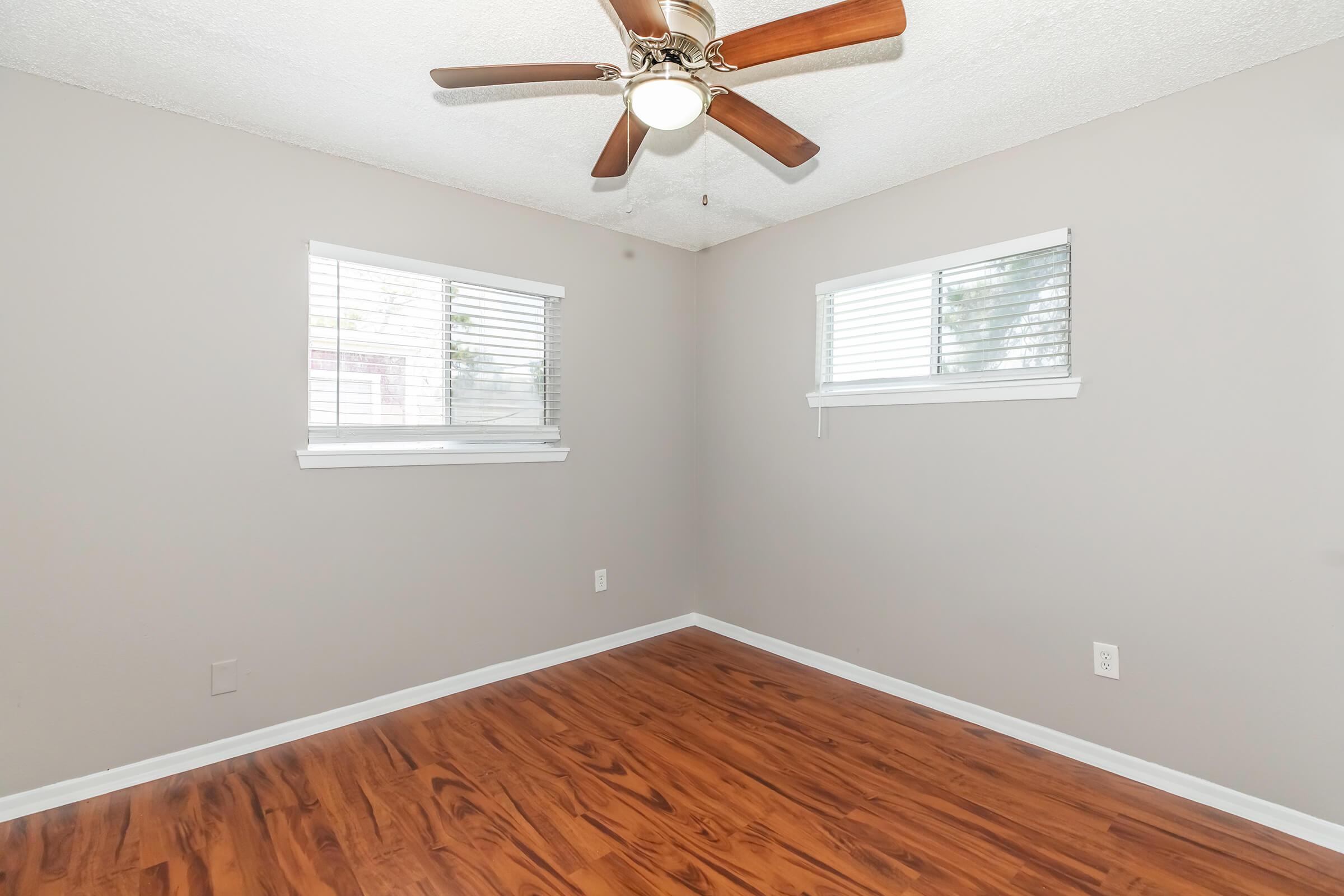 A light gray bedroom with a ceiling fan, two white-framed windows with blinds, and wooden flooring. The walls are bare with no decorations. The overall ambiance is bright and spacious, ideal for personal use or decoration.