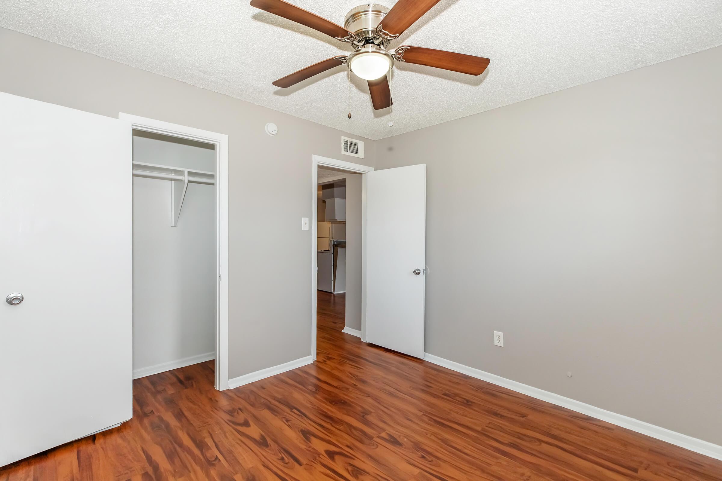 A well-lit, empty room featuring a ceiling fan with wooden blades. The walls are painted a soft gray, and there are two white doors leading to a closet and another room. The floor is made of polished wood, enhancing the warm ambiance of the space.